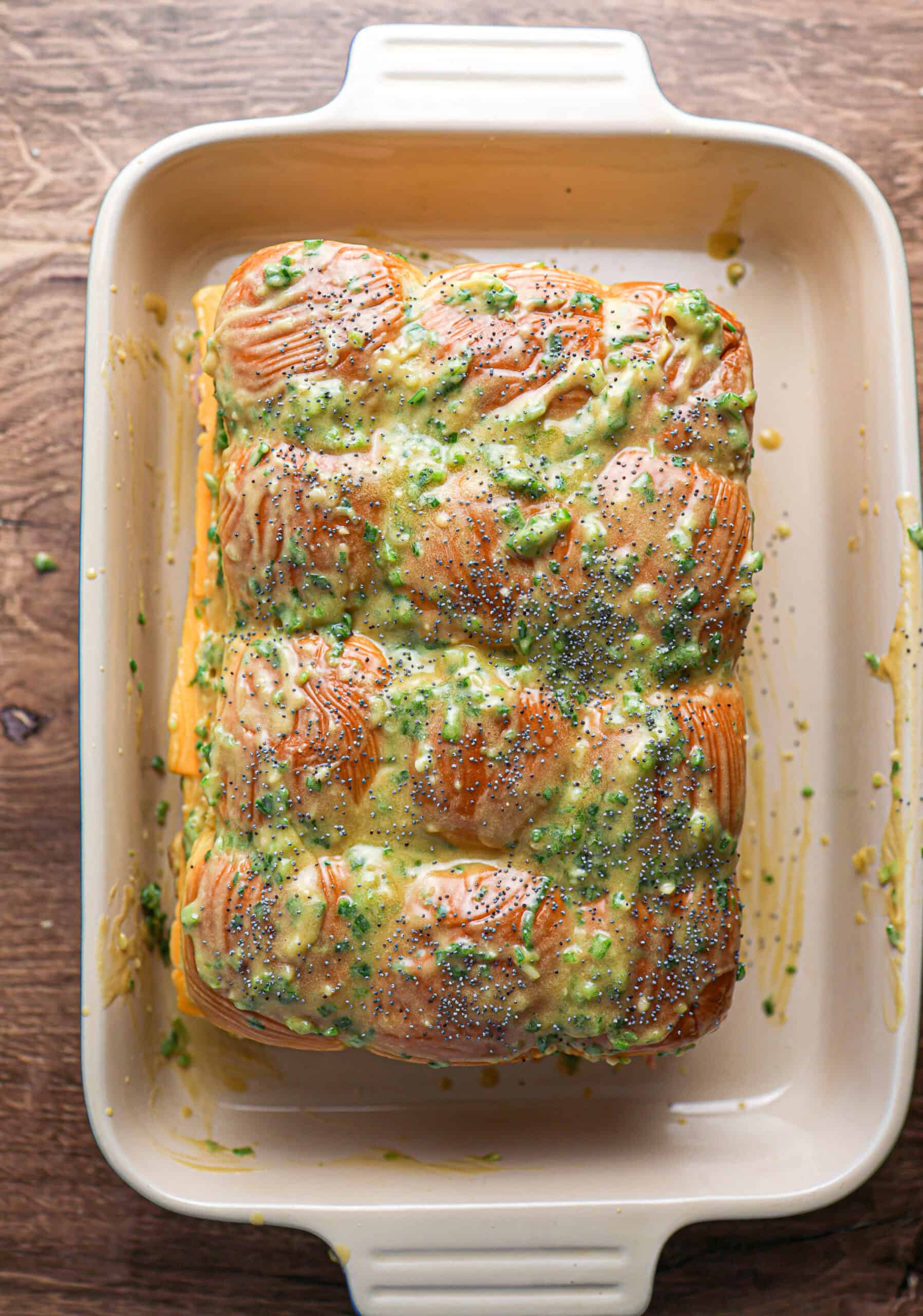 Baked pull-apart bread in a cream-colored dish, inspired by ham and cheese sliders, topped with chopped herbs, poppy seeds, and a yellow sauce or glaze.