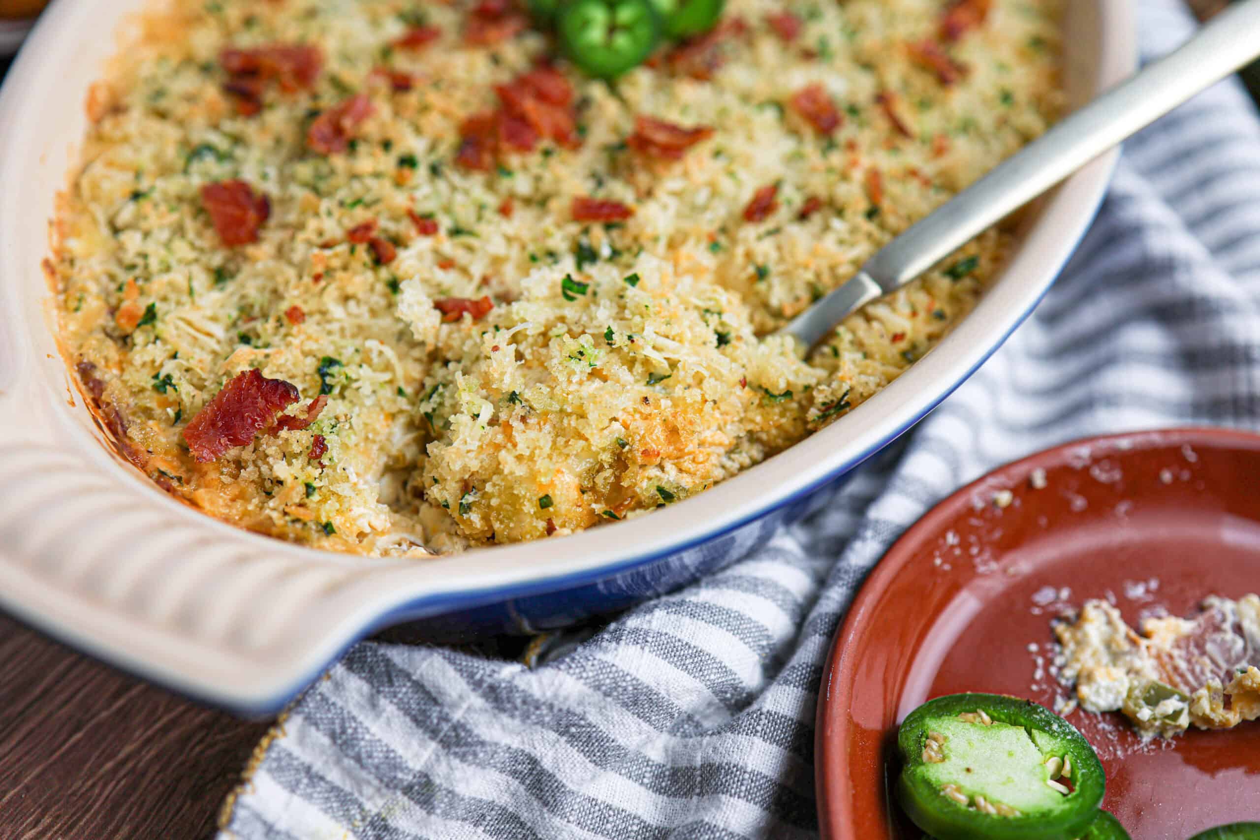 A casserole dish filled with Jalapeño Popper Dip, topped with breadcrumbs, jalapeño slices, and bacon bits, sits on a striped cloth next to a plate with food remnants and extra jalapeño slices.