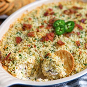 A baked Jalapeño Popper Dip casserole topped with breadcrumbs, chopped bacon, and sliced jalapeños, with a round cracker scooping out a portion. Crackers are visible in the background.