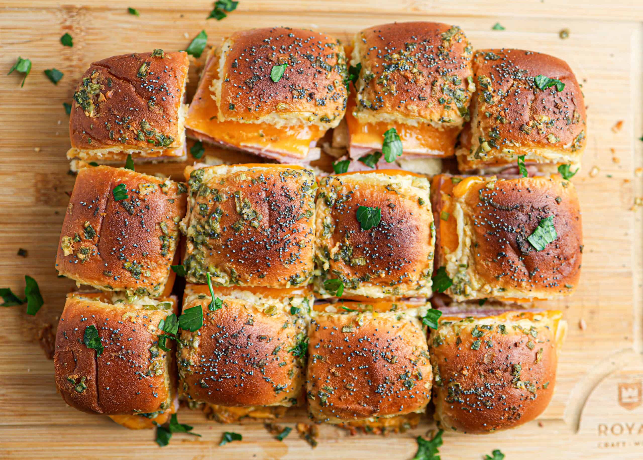 A close-up of twelve ham and cheese sliders on a wooden board, topped with herbs and poppy seeds, and garnished with chopped parsley.