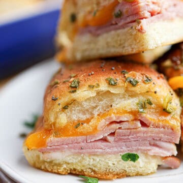 Close-up of ham and cheese sliders on a soft bun, topped with herbs and melted cheddar, served on a white plate.