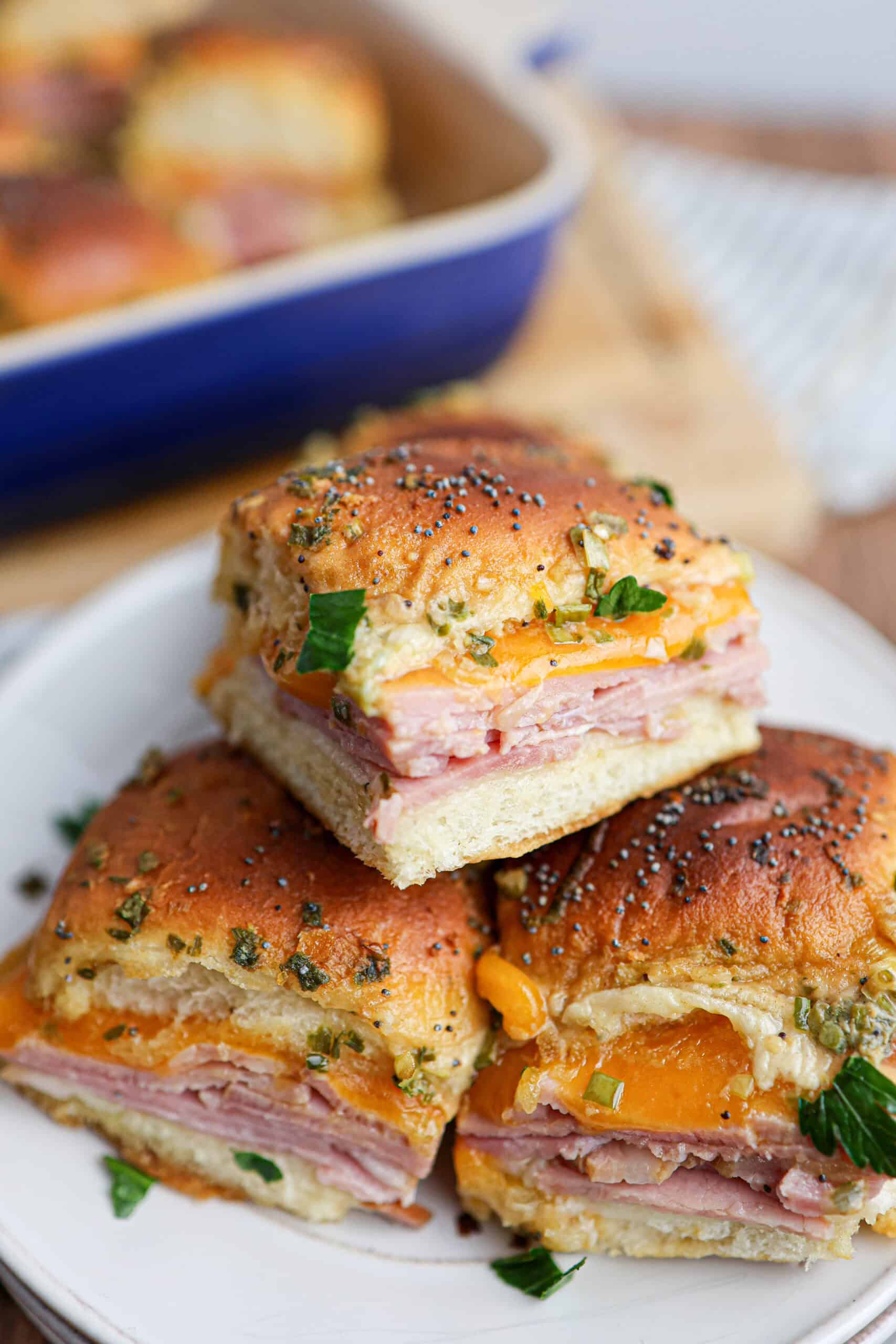 Three ham and cheese sliders with poppy seeds and herbs on top are stacked on a white plate, with a baking dish of freshly baked ham and cheese sliders in the background.