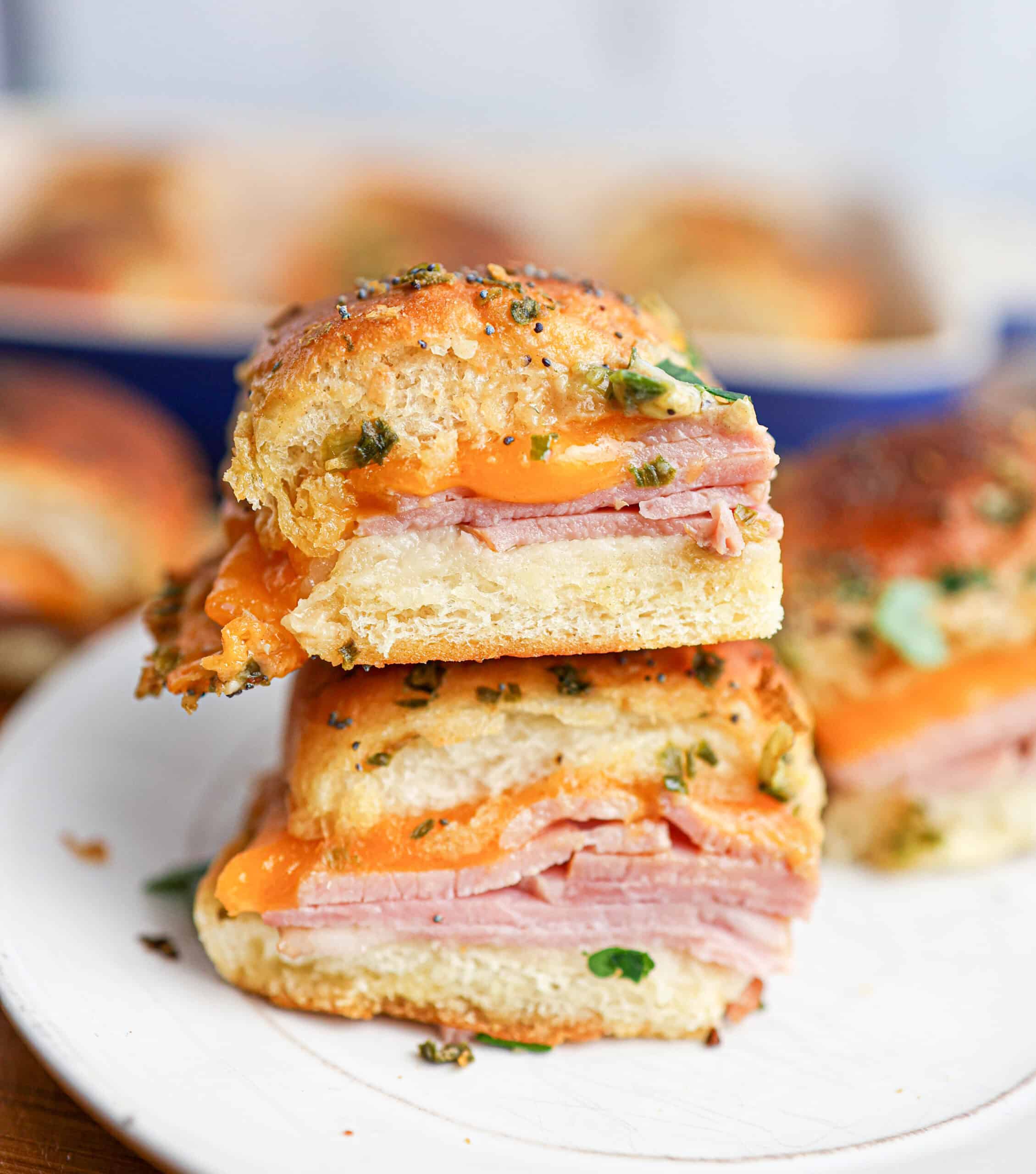 Two ham and cheese sliders stacked on a white plate, with melted cheese and herbs visible on golden, toasted buns—these ham and cheese sliders are perfect for a tasty snack or appetizer.