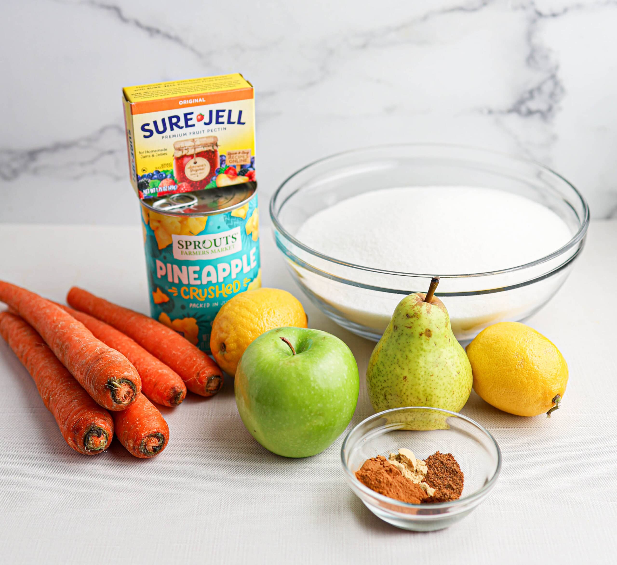 Carrot cake jam ingredients—carrots, green apple, pear, lemons, pineapple, sugar, Sure-Jell pectin, and spices—are arranged on a white surface with a marble background.