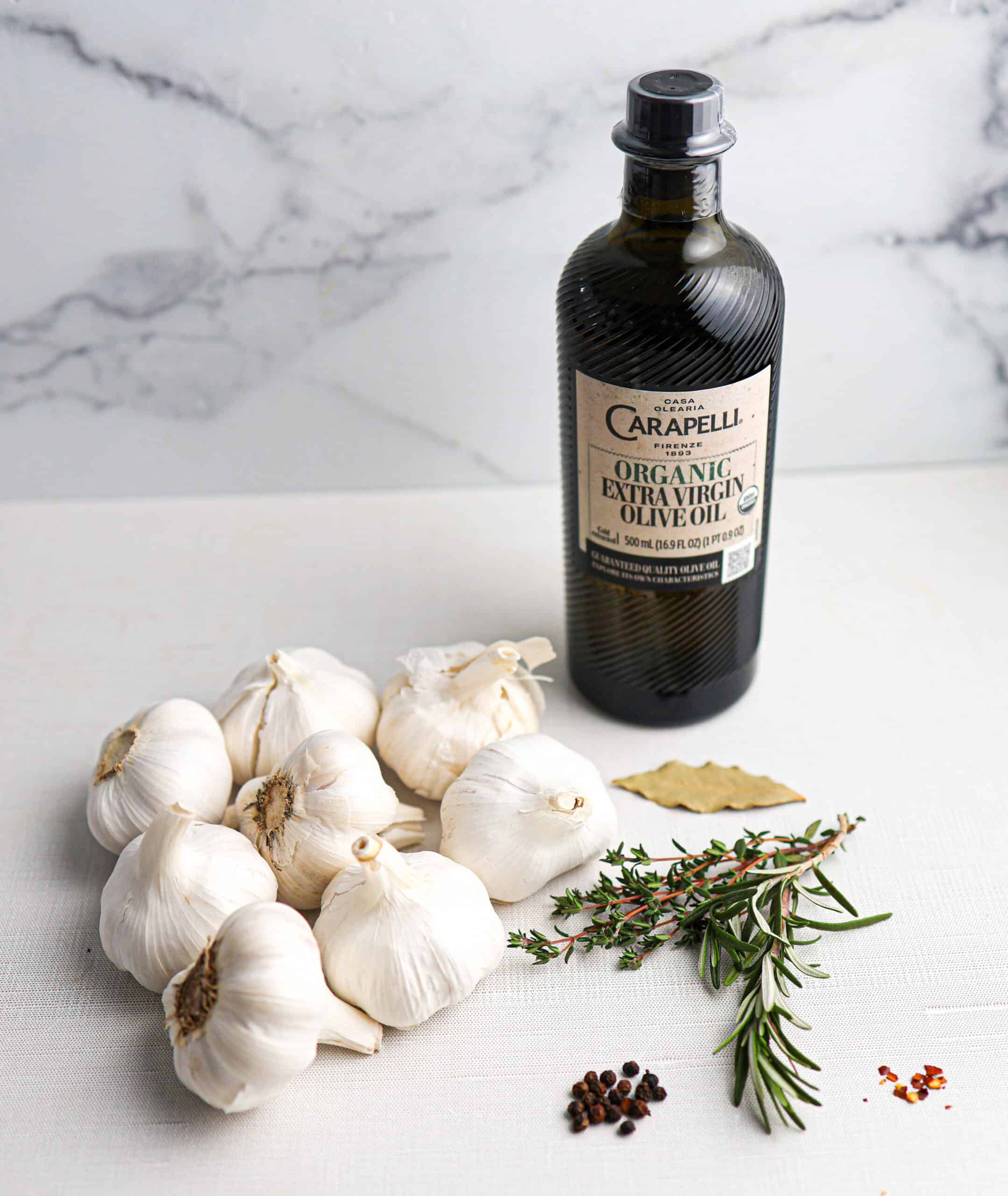 A bottle of Carapelli extra virgin olive oil beside several garlic bulbs, fresh herbs, a bay leaf, peppercorns, and red pepper flakes on a white surface—perfect ingredients for making flavorful garlic confit.