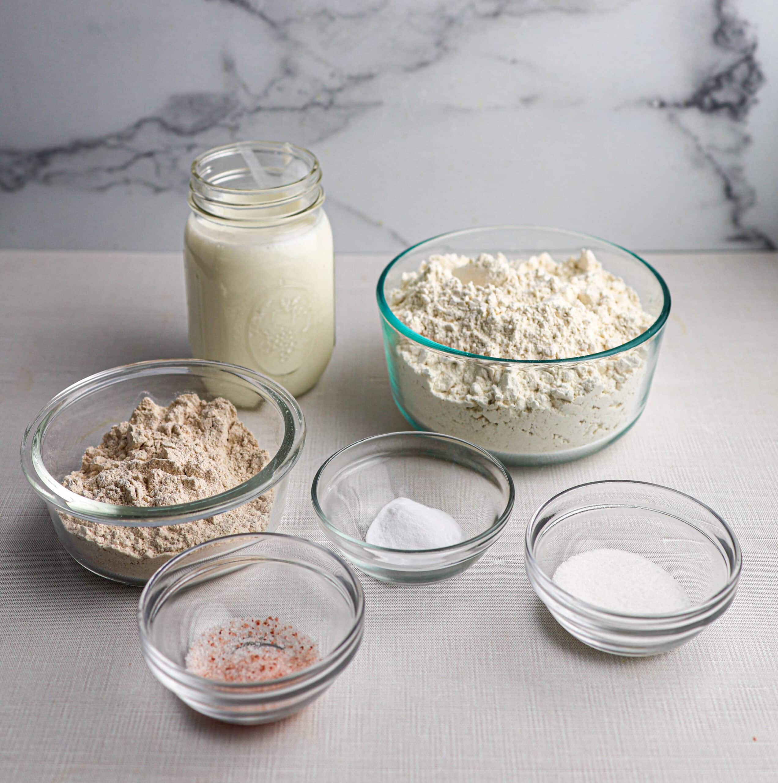 Six glass bowls and a jar display the key ingredients for Irish soda bread—flour, whole wheat flour, baking powder, sugar, salt, and buttermilk arranged on a light surface with a marble background.