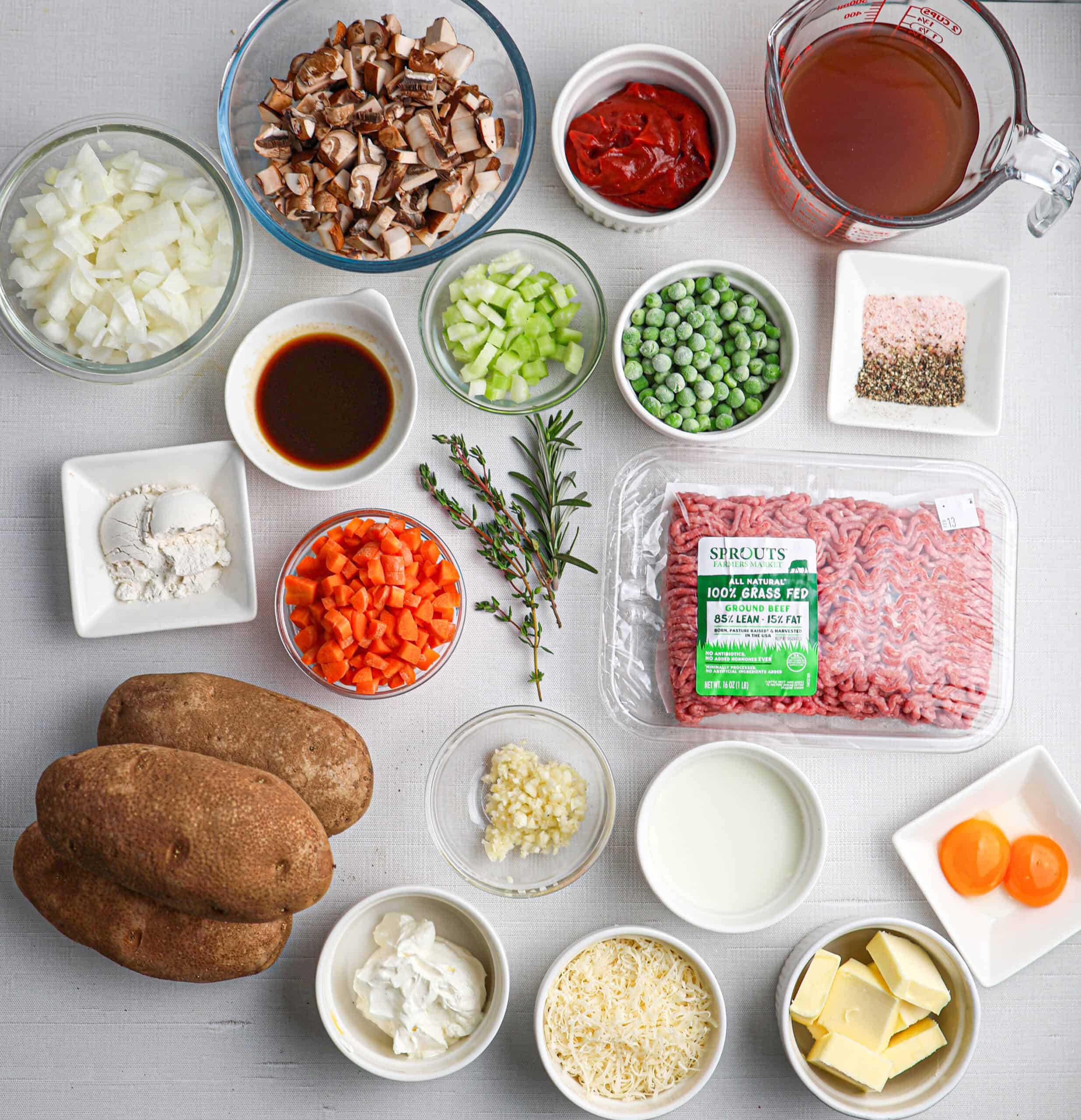 A variety of ingredients for Shepherd's pie, including ground beef, potatoes, chopped vegetables, peas, broth, herbs, seasonings, cheese, flour, milk, butter, and egg yolks on a white surface.