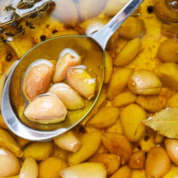 A spoon lifts several cloves of garlic confit from a pot filled with tender garlic, fragrant herbs, and golden oil.