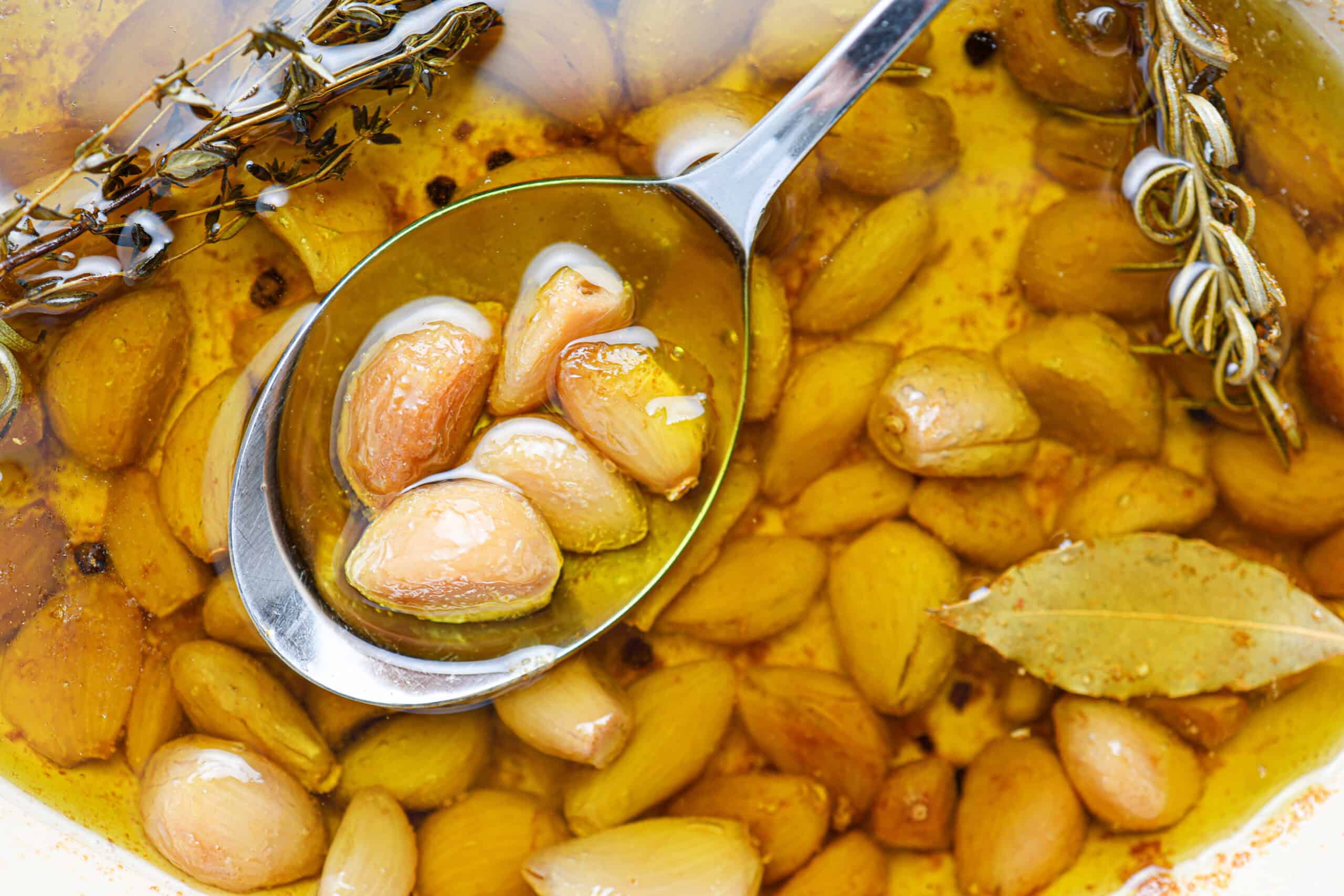 A spoon lifts several cloves of garlic confit from a pot filled with tender garlic, fragrant herbs, and golden oil.