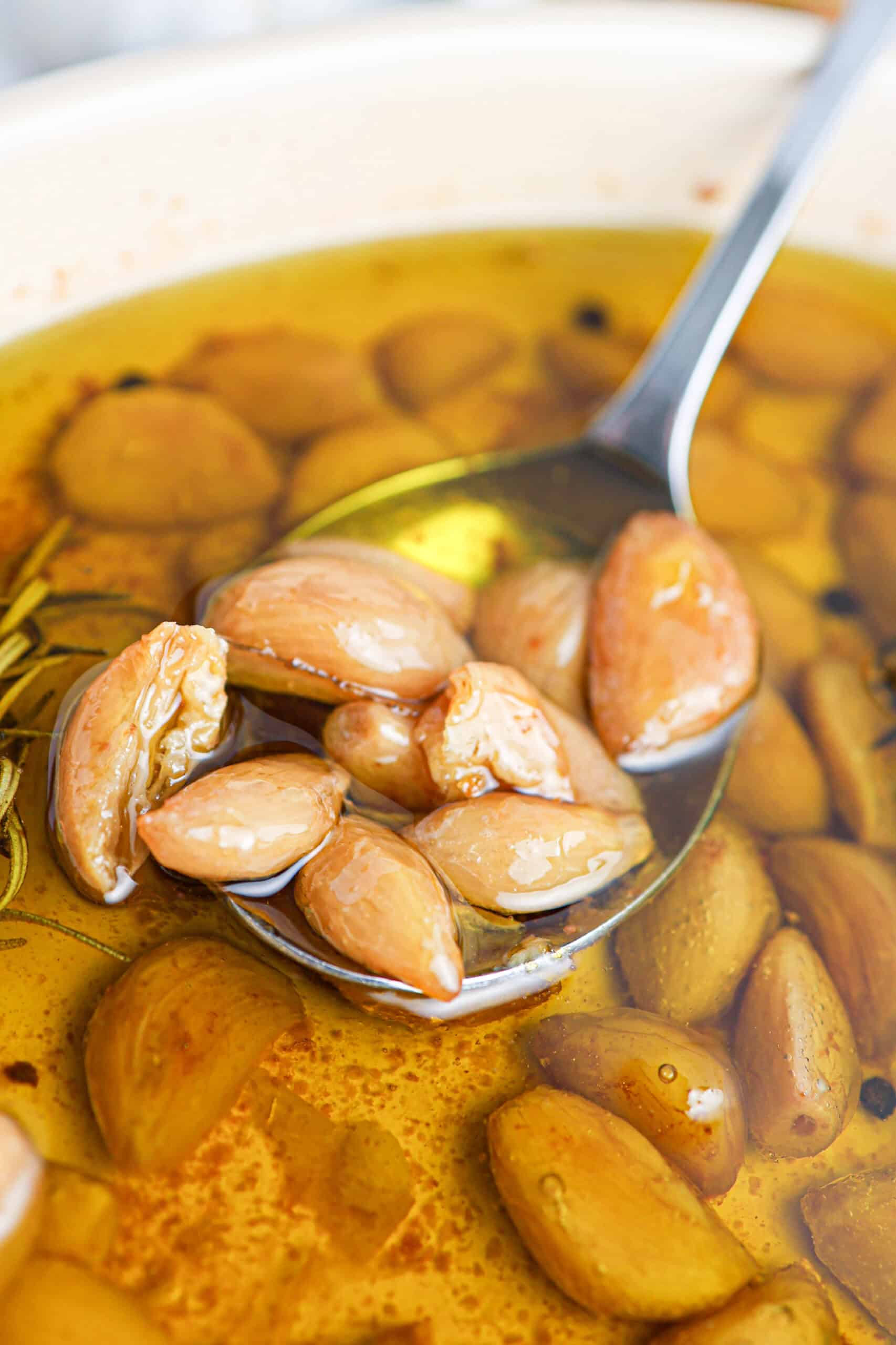 A close-up of a spoon lifting several tender garlic confit cloves from a bowl, each clove glistening as it emerges from the golden oil.