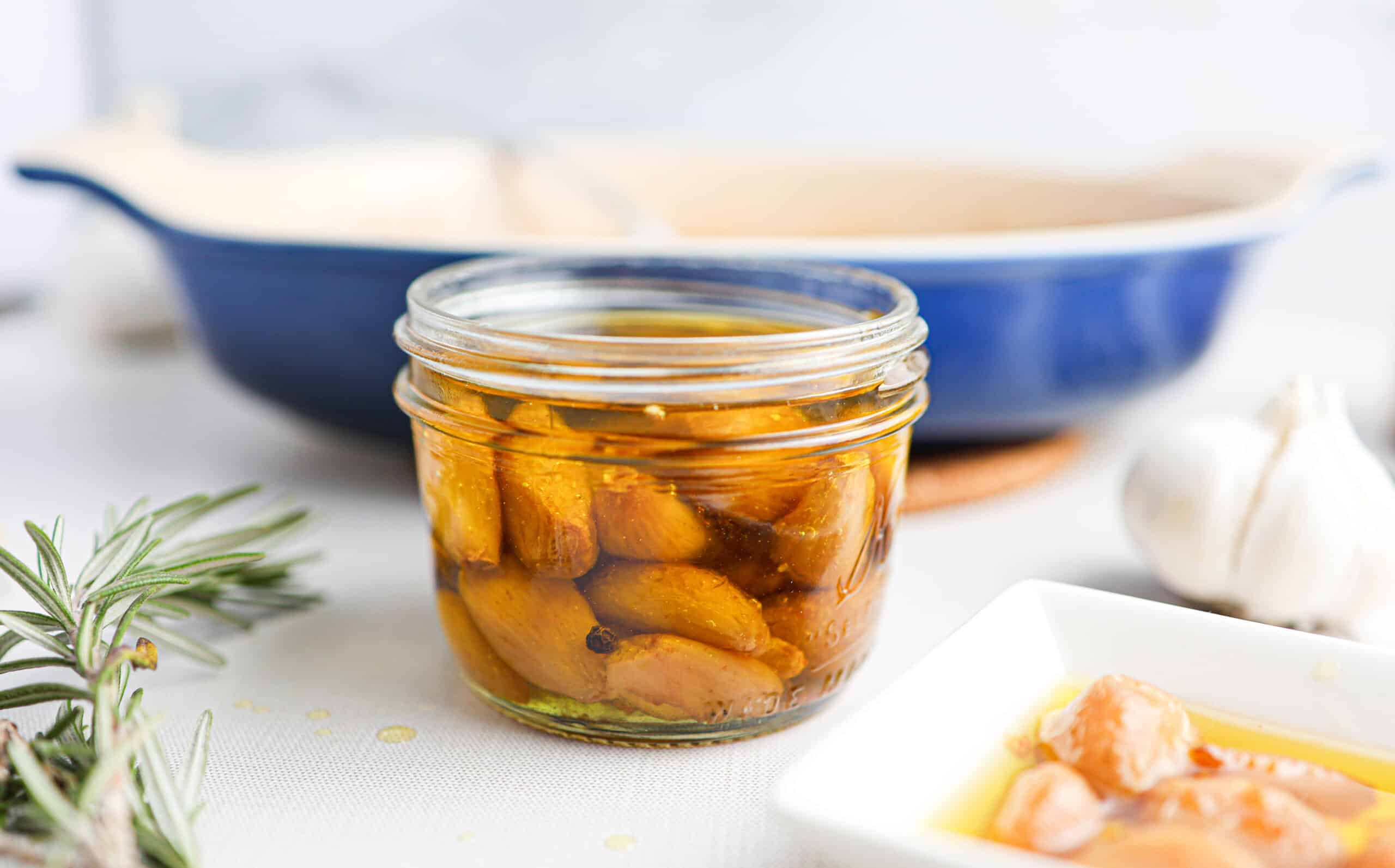 A glass jar filled with garlic confit—tender, roasted cloves in oil—sits on a table, with a blue dish, a garlic bulb, and a rosemary sprig in the background.
