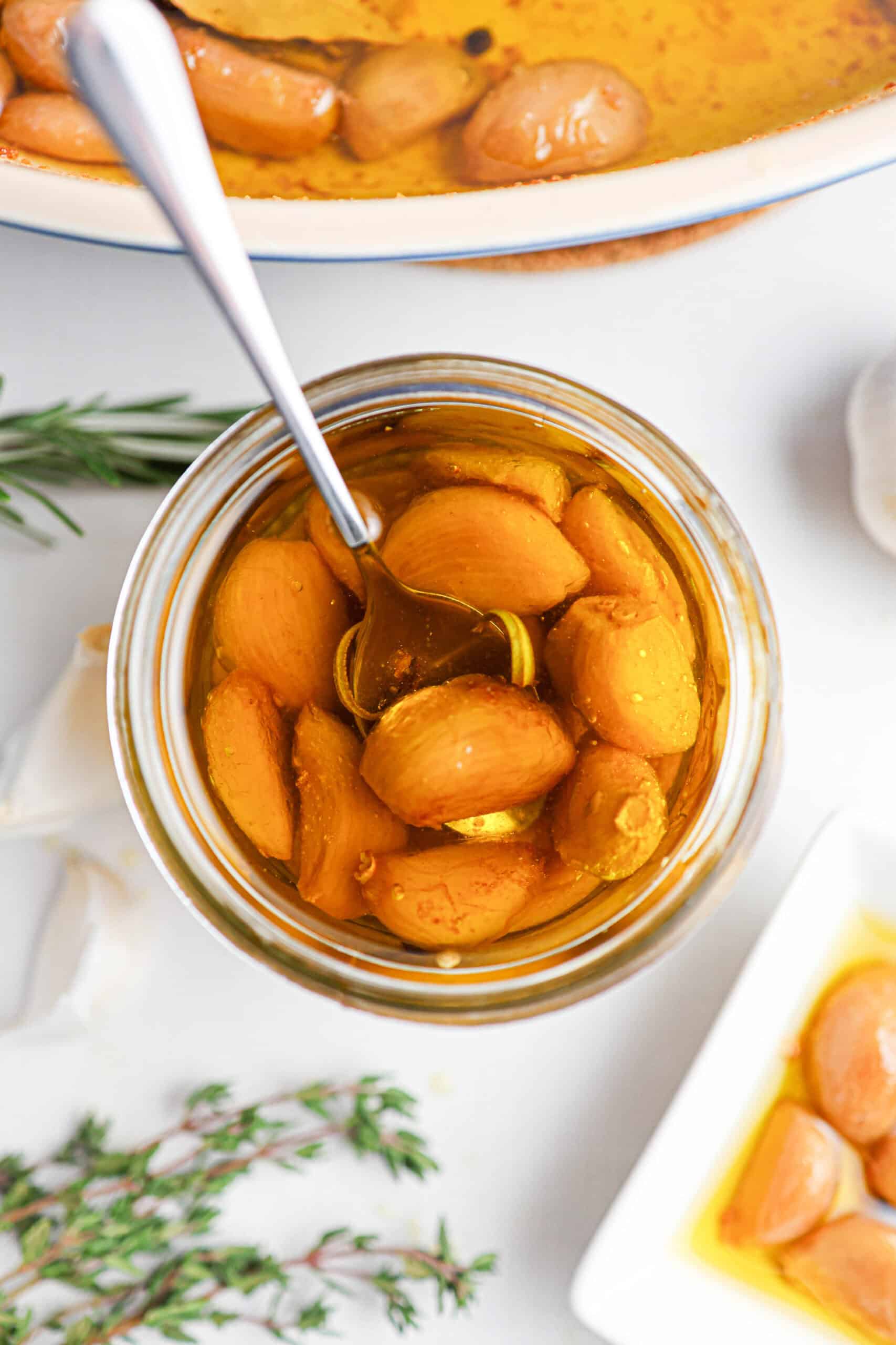 A jar of garlic confit—garlic cloves submerged in oil, with a spoon inside. Fresh herbs and a dish with some confit cloves sit nearby on a white surface.