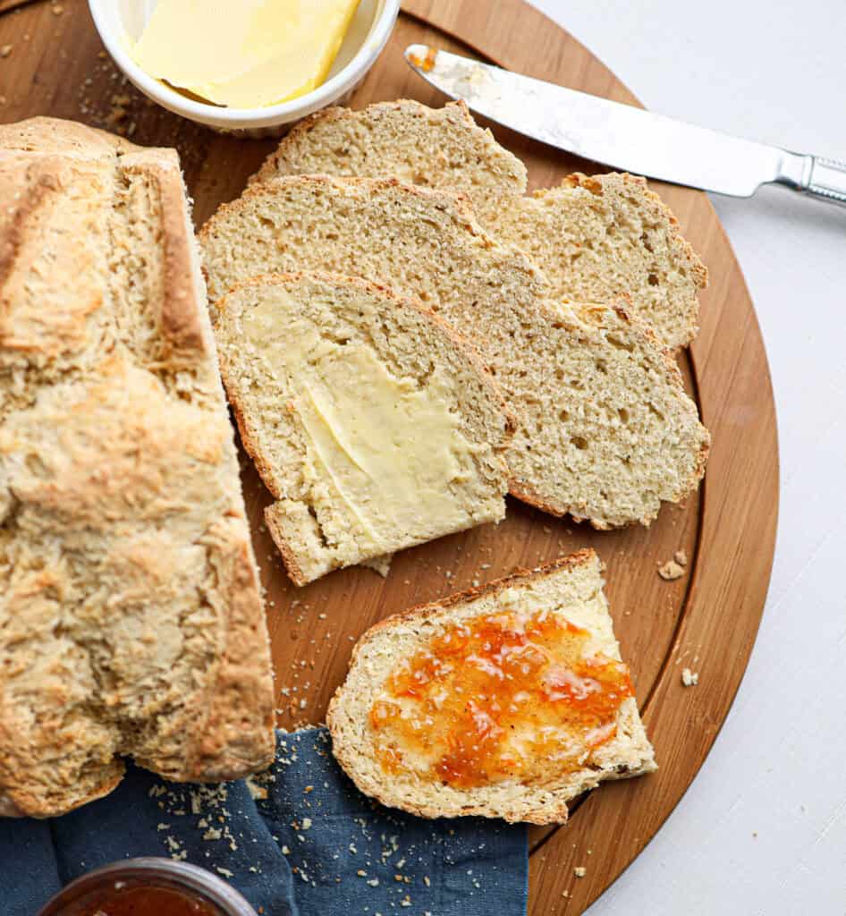 A round wooden board with sliced Irish soda bread, one slice spread with butter and another with orange marmalade; a butter knife and dish are nearby.