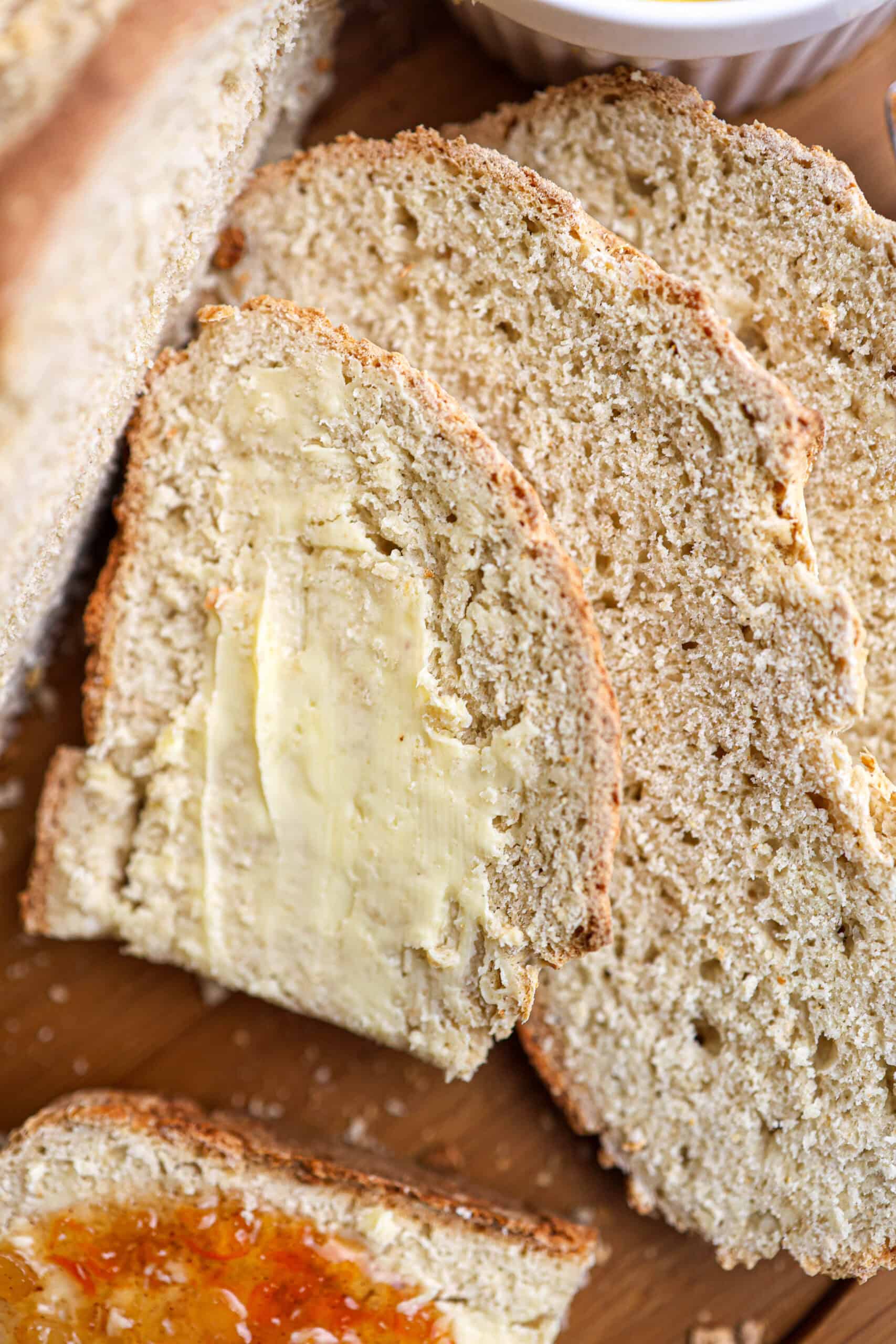 Three slices of Irish soda bread rest on a wooden surface, one spread with butter and another with orange jam.