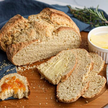 A loaf of sliced Irish soda bread sits on a wooden board, accompanied by butter, a butter knife, jam, and a sprig of rosemary nearby.