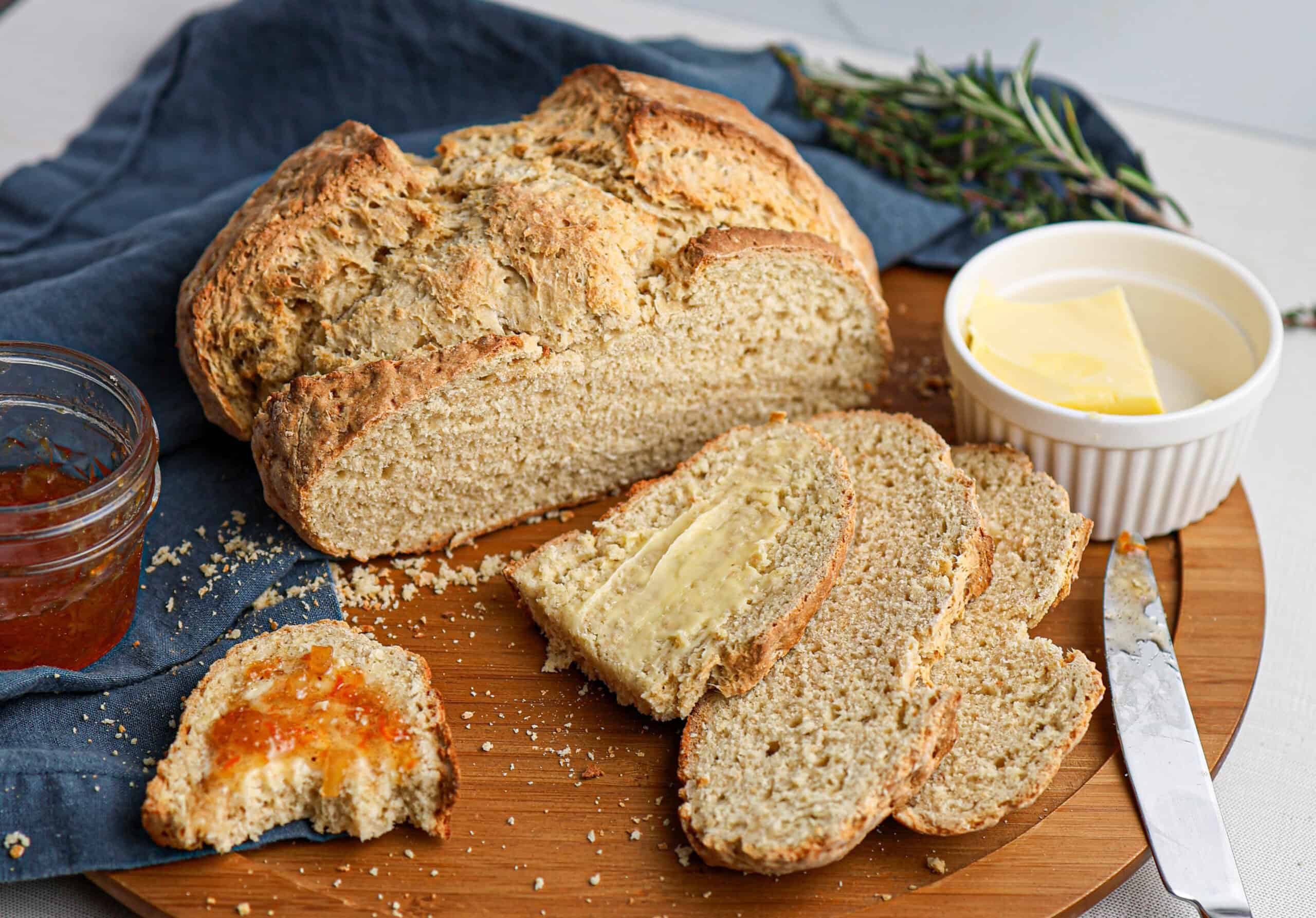 A loaf of sliced Irish soda bread sits on a wooden board, accompanied by butter, a butter knife, jam, and a sprig of rosemary nearby.