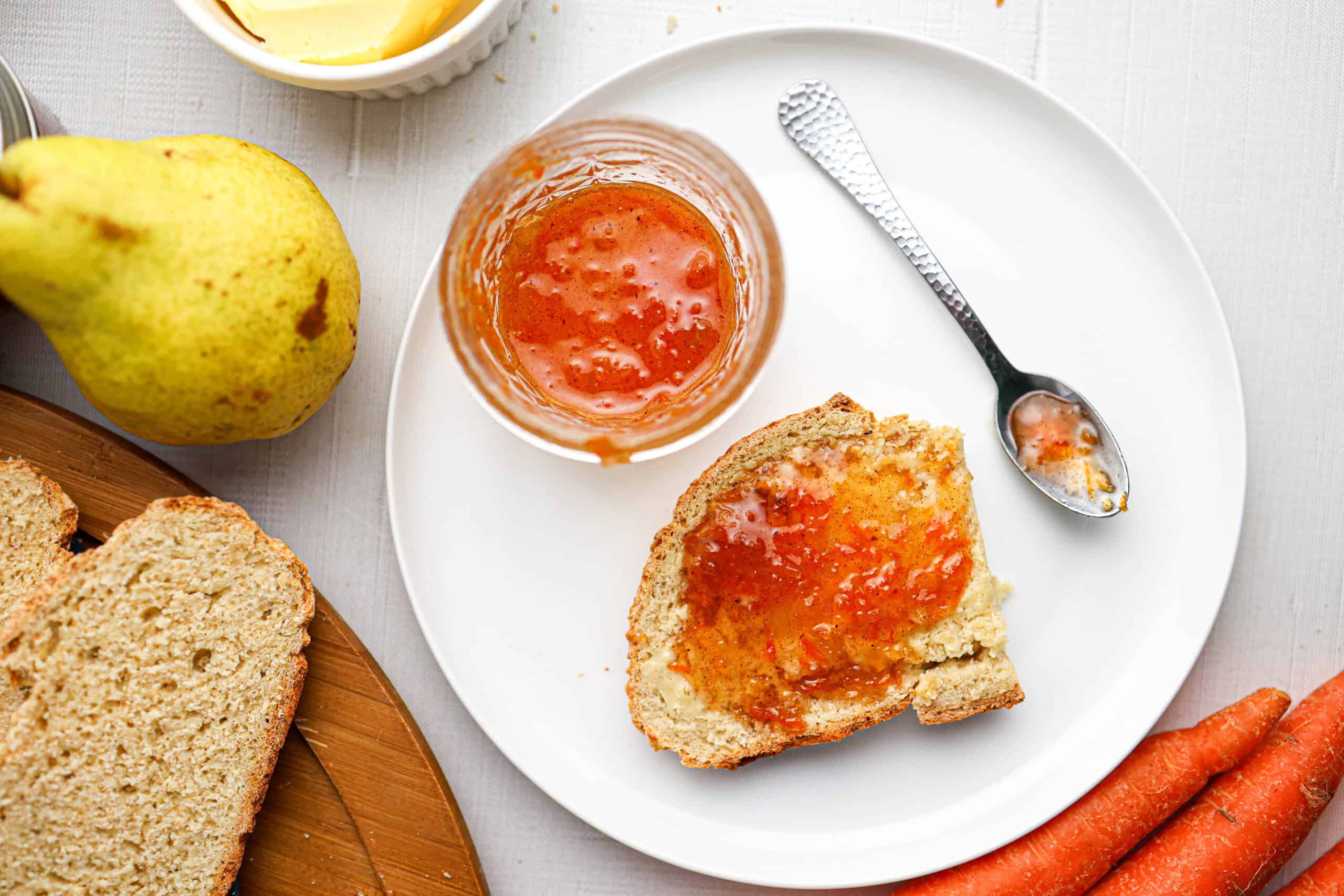 A plate with a slice of bread spread with carrot cake jam, a jar of jam with a spoon, a pear, sliced bread, and carrots on a white surface.