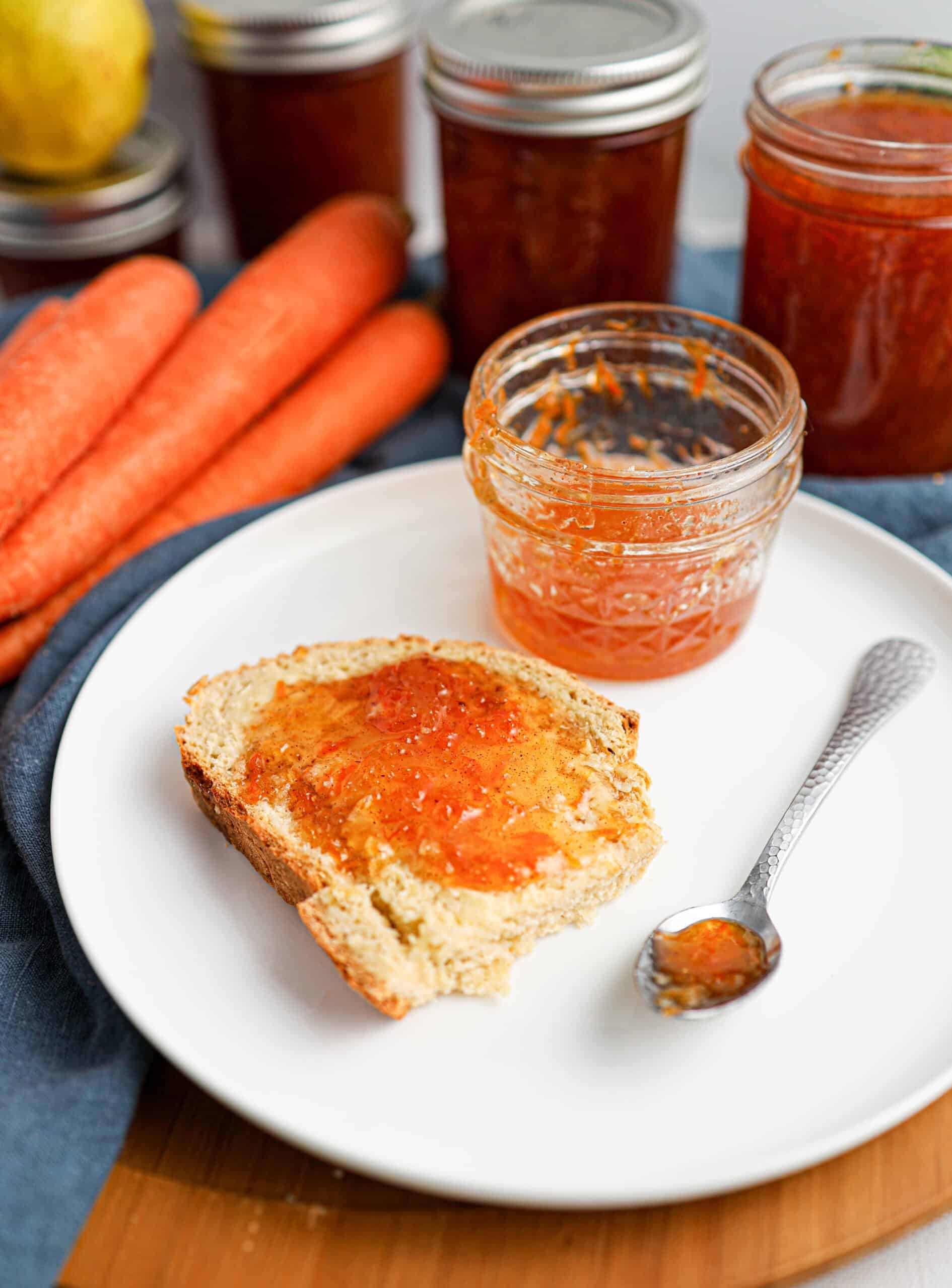 A slice of bread with carrot cake jam sits on a white plate next to a jar of jam and a spoon, with fresh carrots and more jam jars in the background.