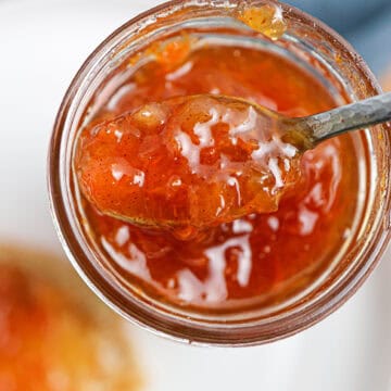 A close-up of a spoon scooping carrot cake jam from a glass jar, with the jam's glossy, orange-colored texture visible.