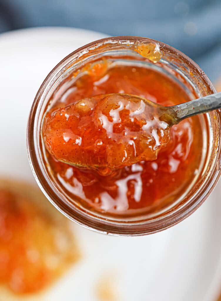 A close-up of a spoon scooping carrot jam from a glass jar, with the jam's glossy, orange-colored texture visible.