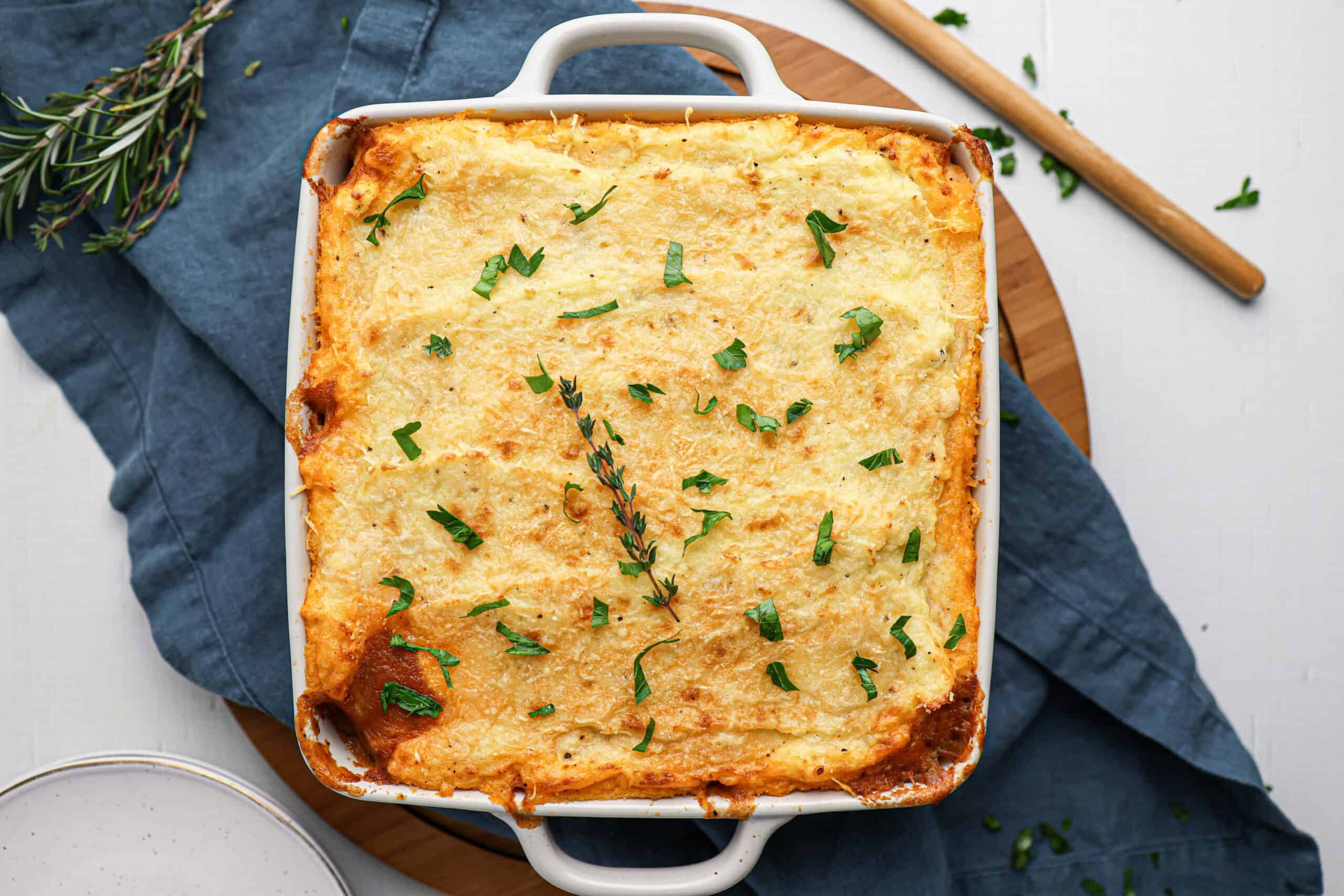 A classic Shepherd's pie casserole topped with melted cheese and garnished with chopped herbs, set on a blue cloth with a sprig of rosemary nearby.
