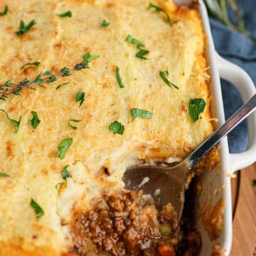 A classic Shepherd's pie in a white dish with a serving spoon, featuring a golden layer of mashed potatoes atop savory ground meat and vegetable filling, garnished with fresh herbs.