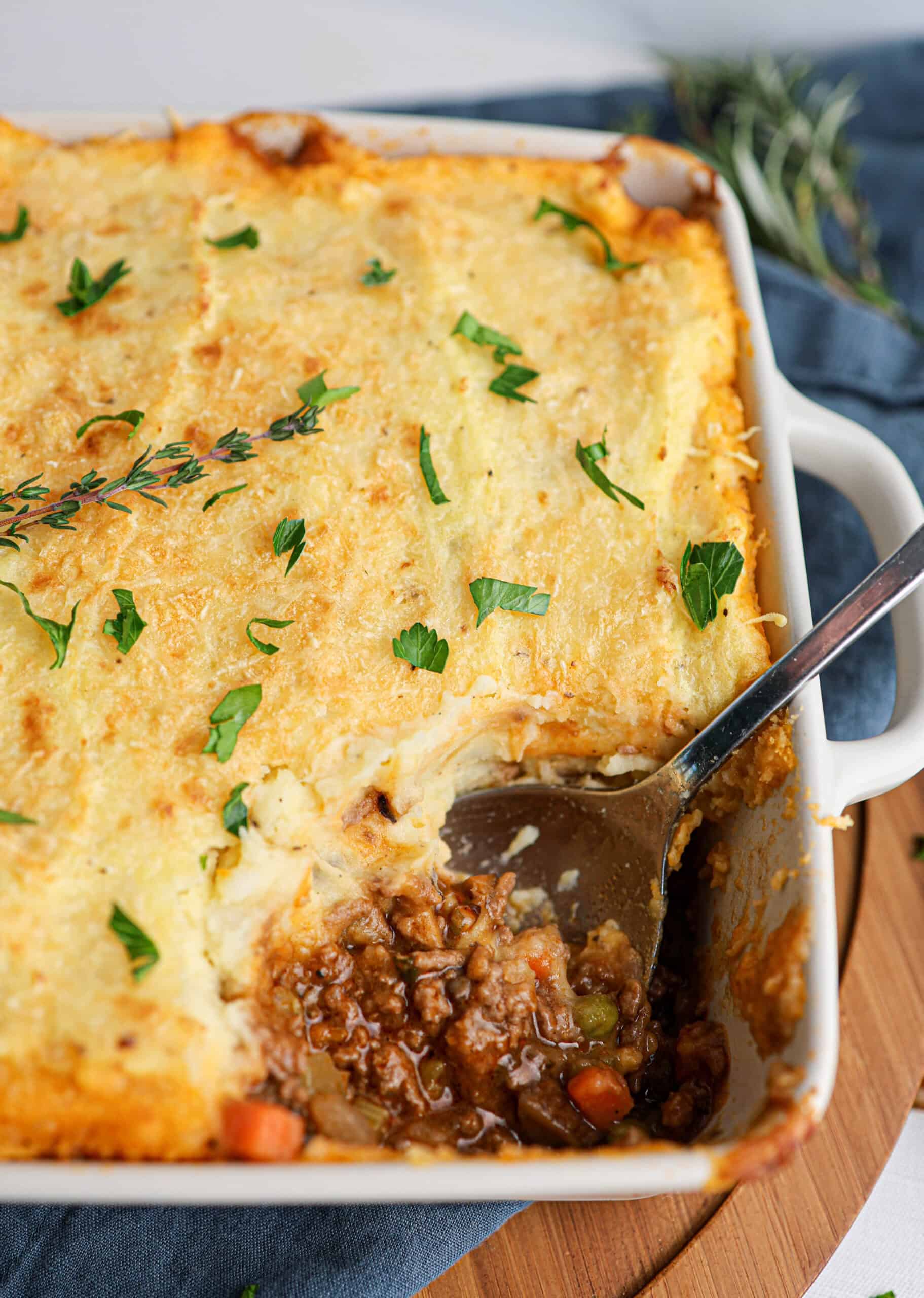 A classic Shepherd's pie in a white dish with a serving spoon, featuring a golden layer of mashed potatoes atop savory ground meat and vegetable filling, garnished with fresh herbs.