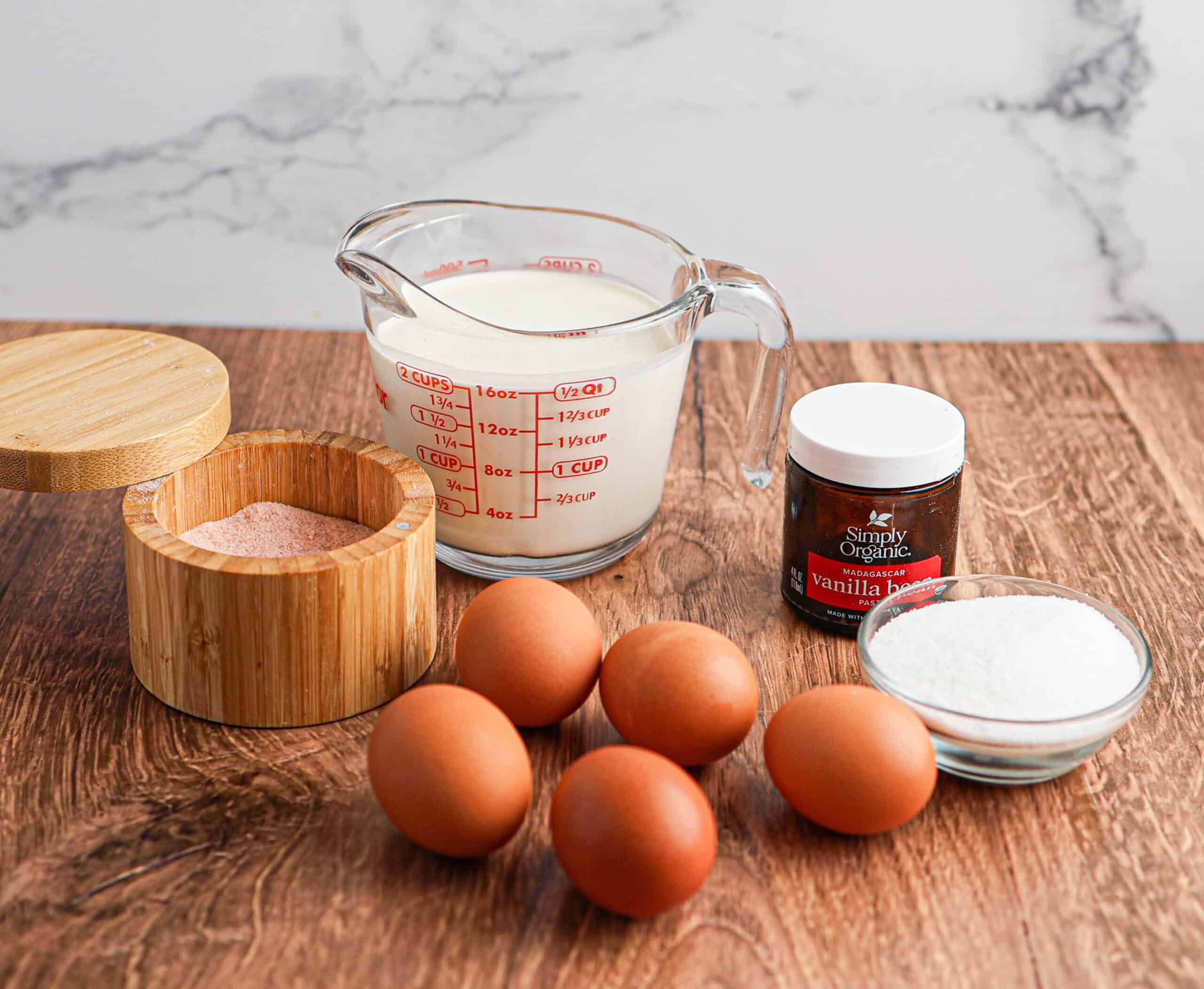 Five eggs, a glass measuring cup of milk, a jar of vanilla bean paste, a bowl of sugar, and a wooden salt container lay ready on the wood surface—perfect ingredients for making classic Crème Brûlée.