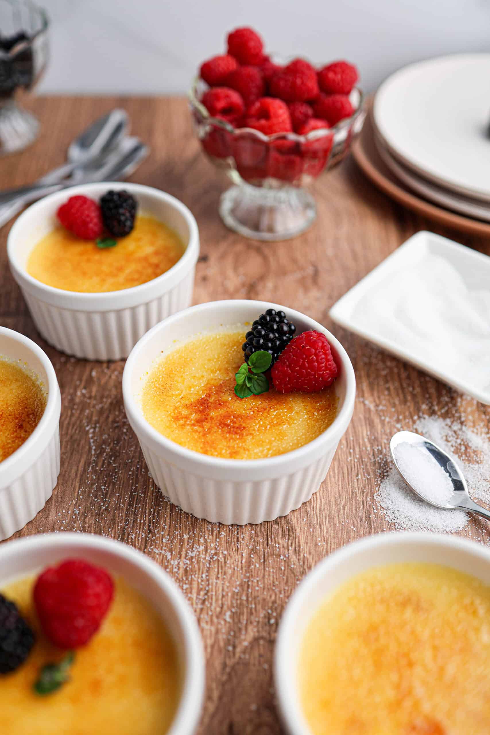 Individual servings of the best homemade Crème Brûlée in white ramekins, each topped with a raspberry, a blackberry, and a mint leaf, with a bowl of raspberries and plates in the background.