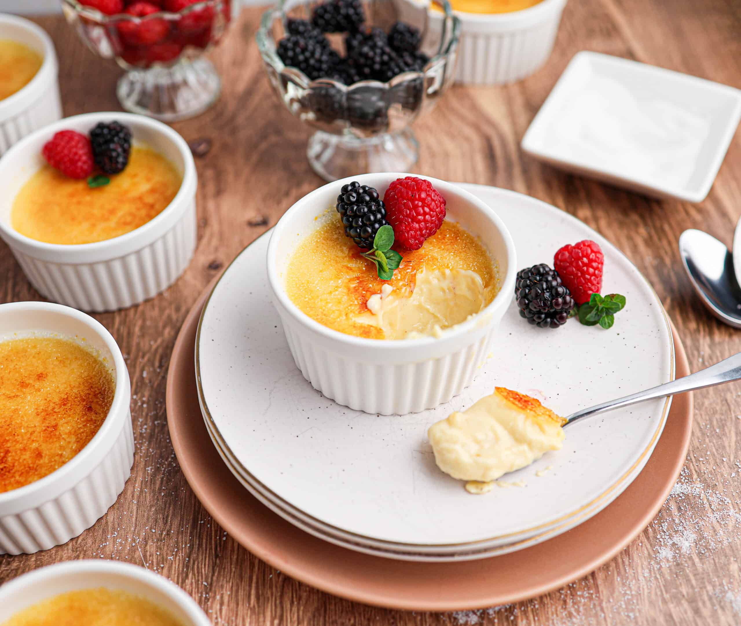 Several ramekins of classic Crème Brûlée are topped with fresh raspberries and blackberries on a wooden table, with one ramekin partially eaten and a spoonful served on a plate.