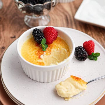 A ramekin of classic Crème Brûlée topped with blackberries, raspberries, and mint sits on a plate, with a spoonful of silky custard resting beside it.