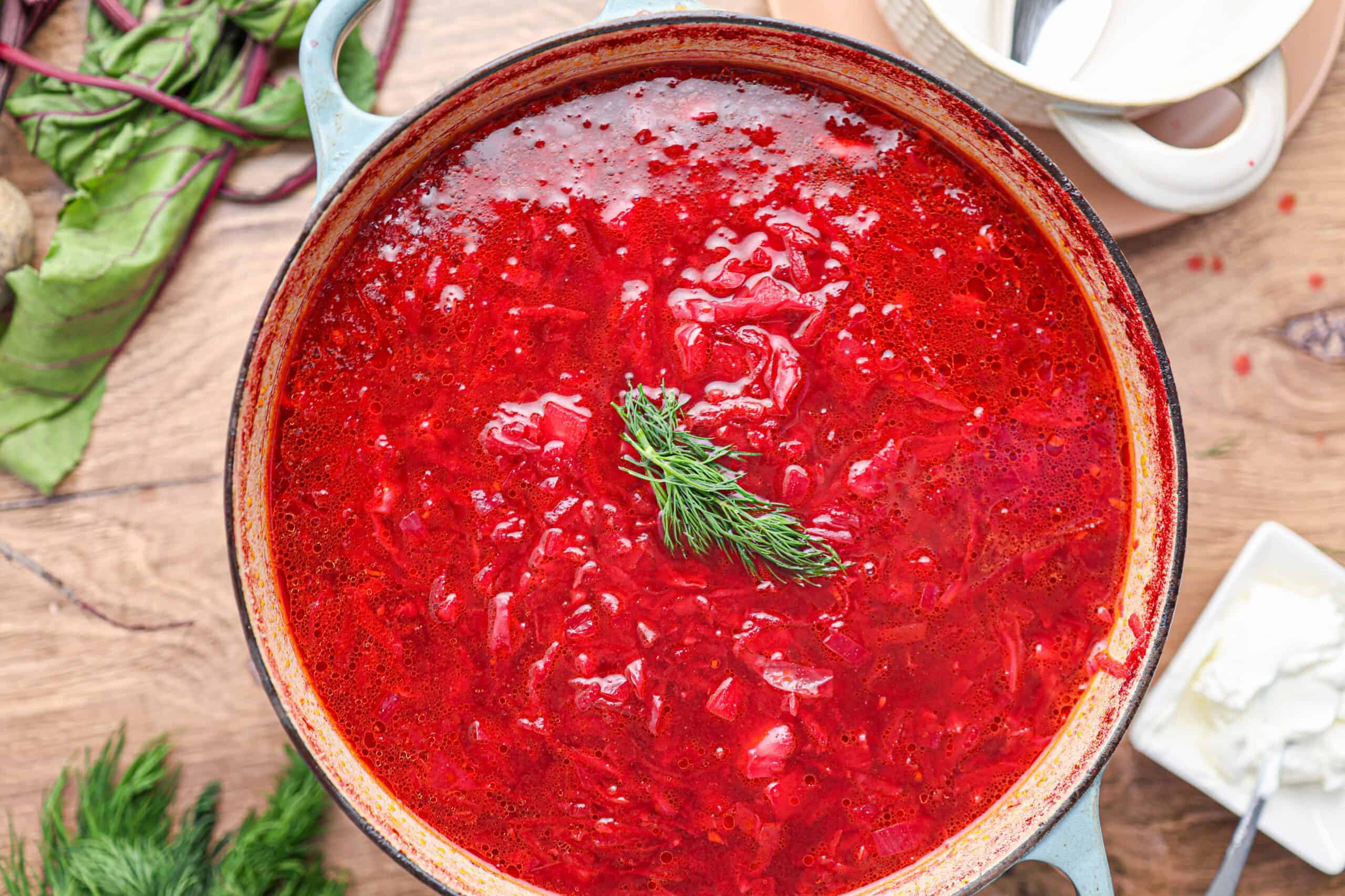 A pot of vibrant borscht, garnished with a sprig of fresh dill, is viewed from above on a wooden table.
