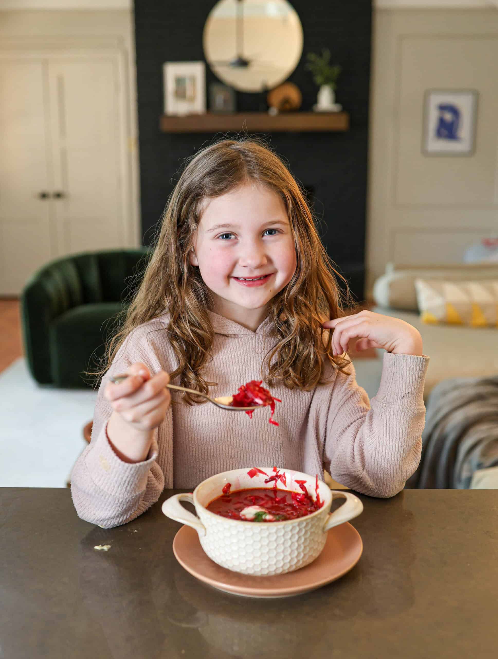 A young girl with long hair sits at a table, smiling and holding a spoonful of beet soup above a bowl in a cozy, modern living room.