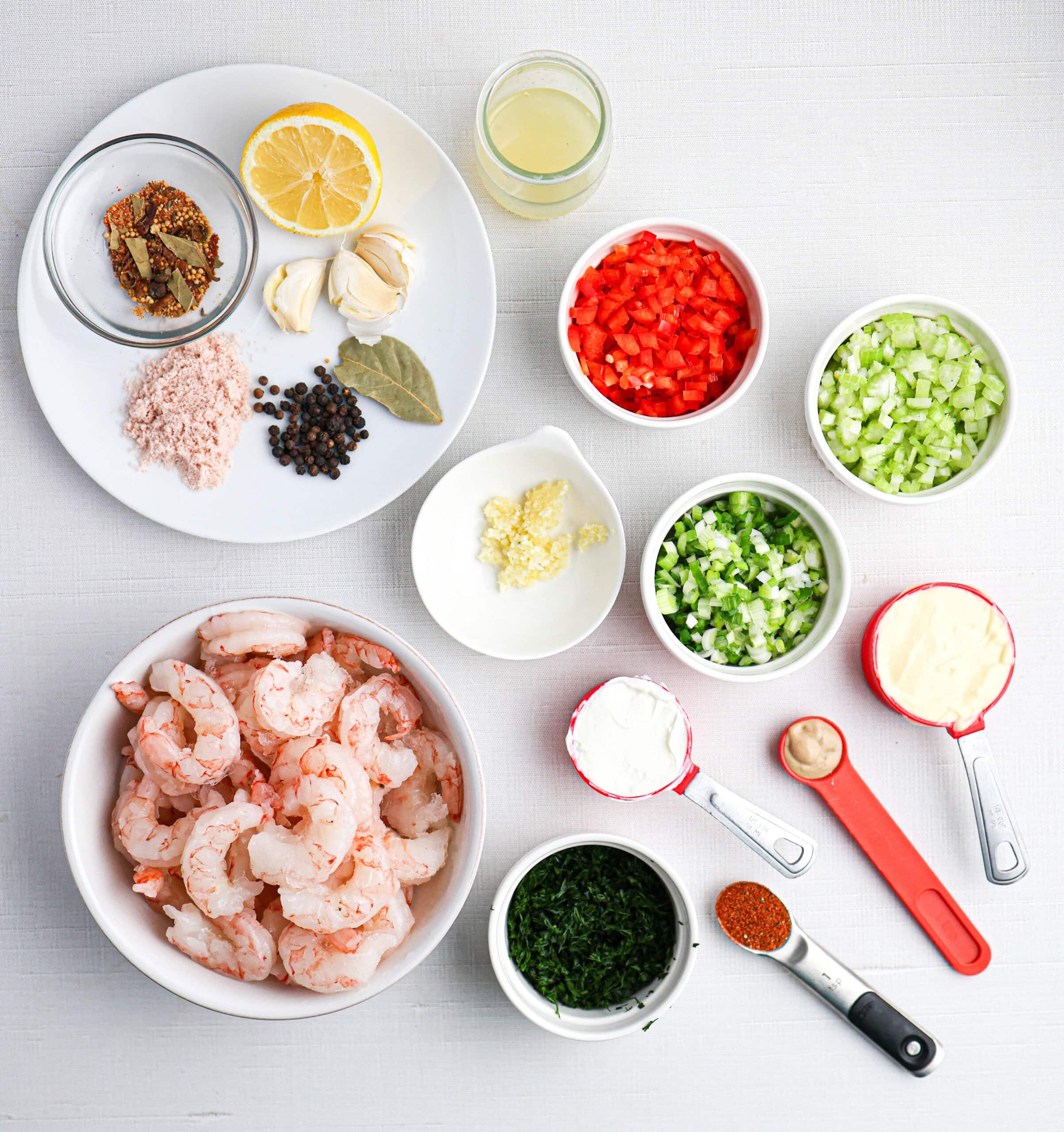 Ingredients for a shrimp salad dish arranged on a white surface, including shrimp, spices, fresh vegetables, lemon, fresh herbs, mayonnaise, and seasonings in bowls and measuring spoons.