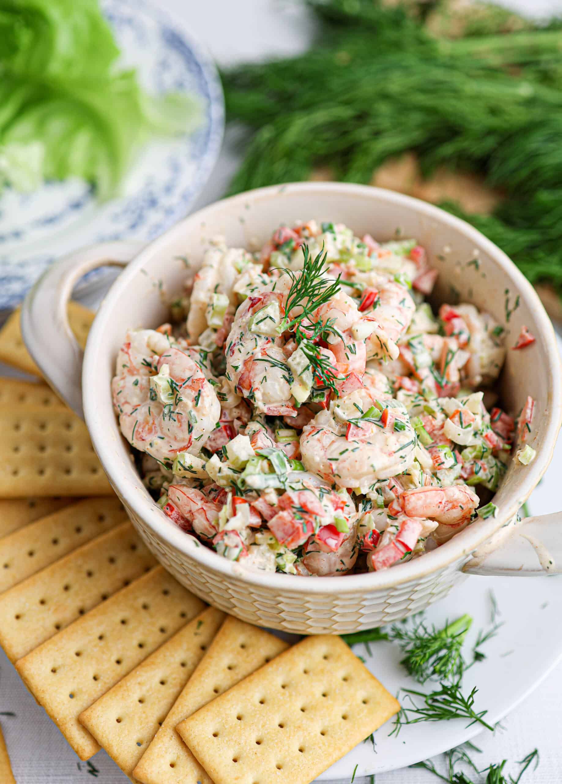 A bowl of creamy shrimp salad with herbs, garnished with fresh dill, surrounded by rectangular crackers on a white surface.