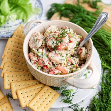 A bowl of shrimp salad with herbs and vegetables is served with a spoon, surrounded by crackers, fresh dill, and crisp lettuce on a table.