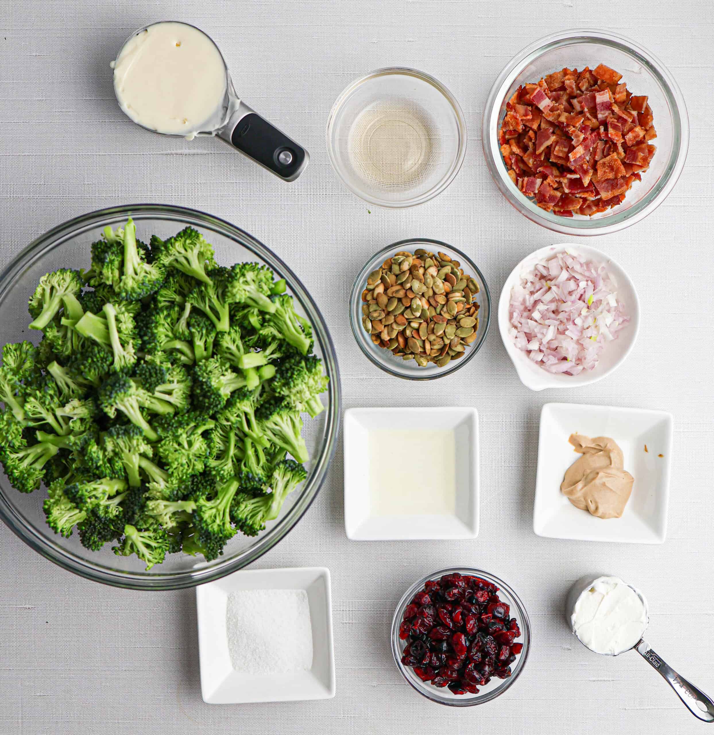 Nine bowls and dishes containing ingredients for a classic broccoli bacon salad: broccoli florets, bacon bits, pumpkin seeds, diced shallots, dried cranberries, mayonnaise, vinegar, sugar, Dijon mustard, and cream cheese on a white surface.