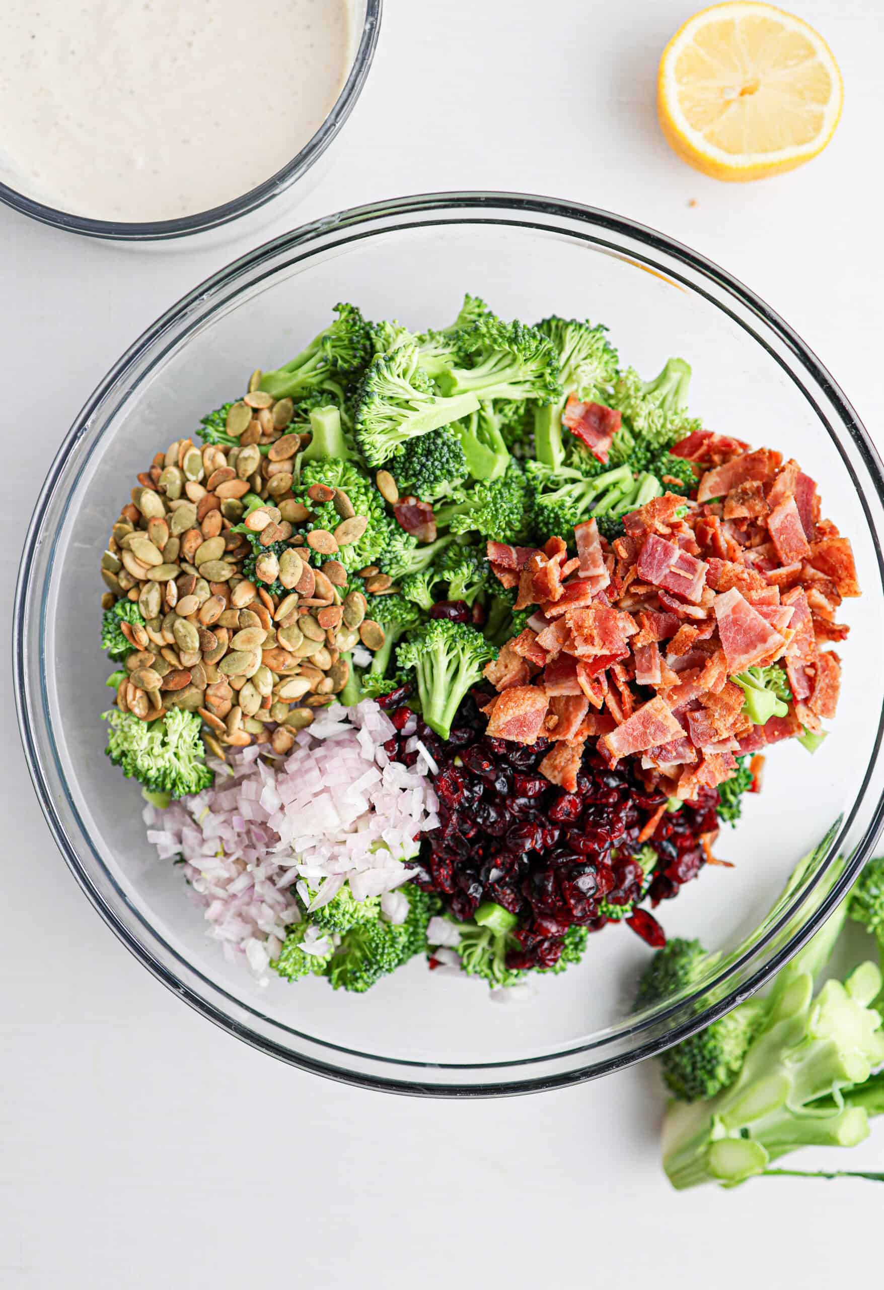 A glass bowl containing broccoli bacon salad made with chopped broccoli, cooked bacon, pumpkin seeds, diced red onion, and dried cranberries, with a half lemon, broccoli florets, and a bowl of dressing nearby.