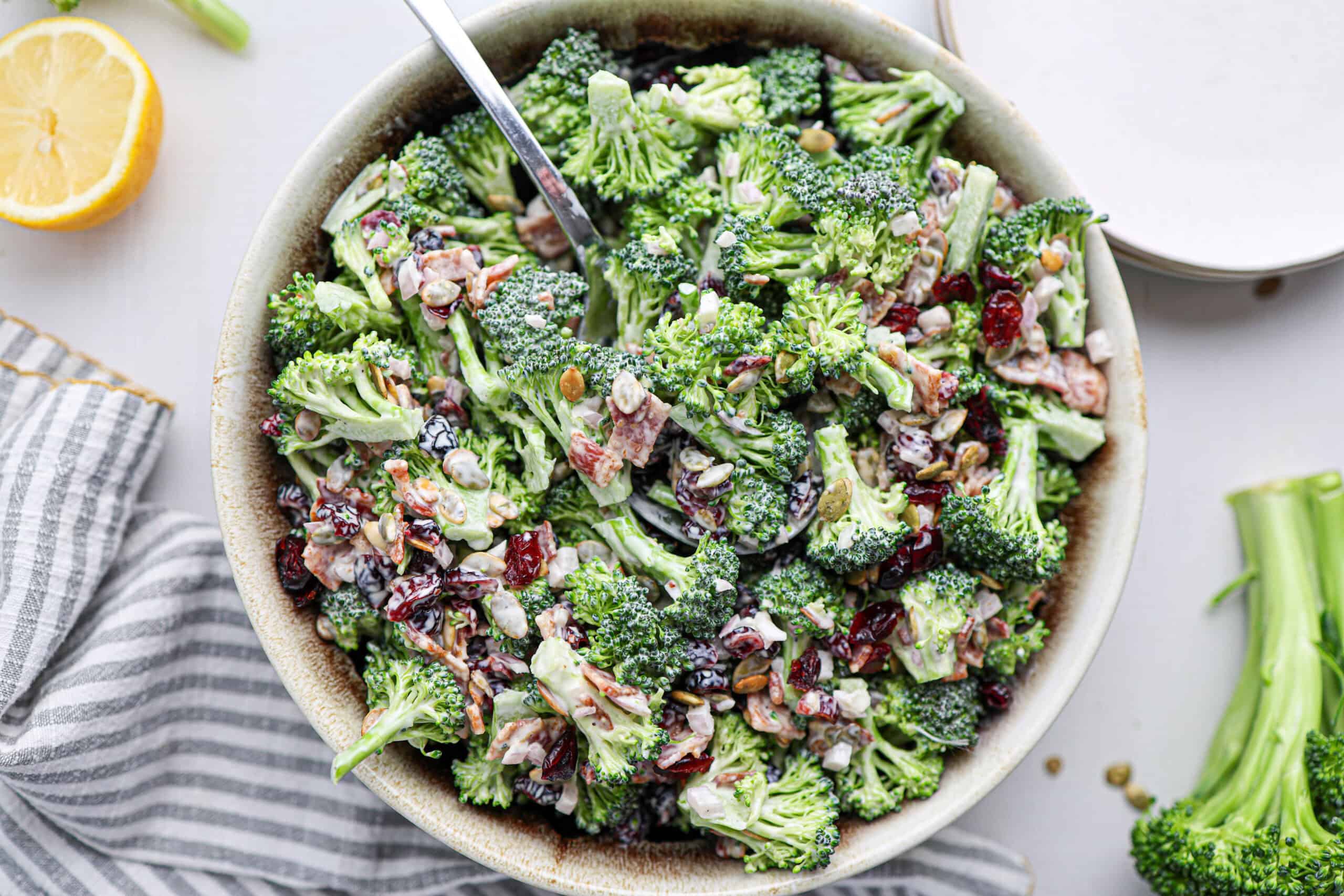 A bowl of broccoli bacon salad with dried cranberries, sunflower seeds, crispy bacon pieces, and a creamy dressing, served with a spoon in the bowl.