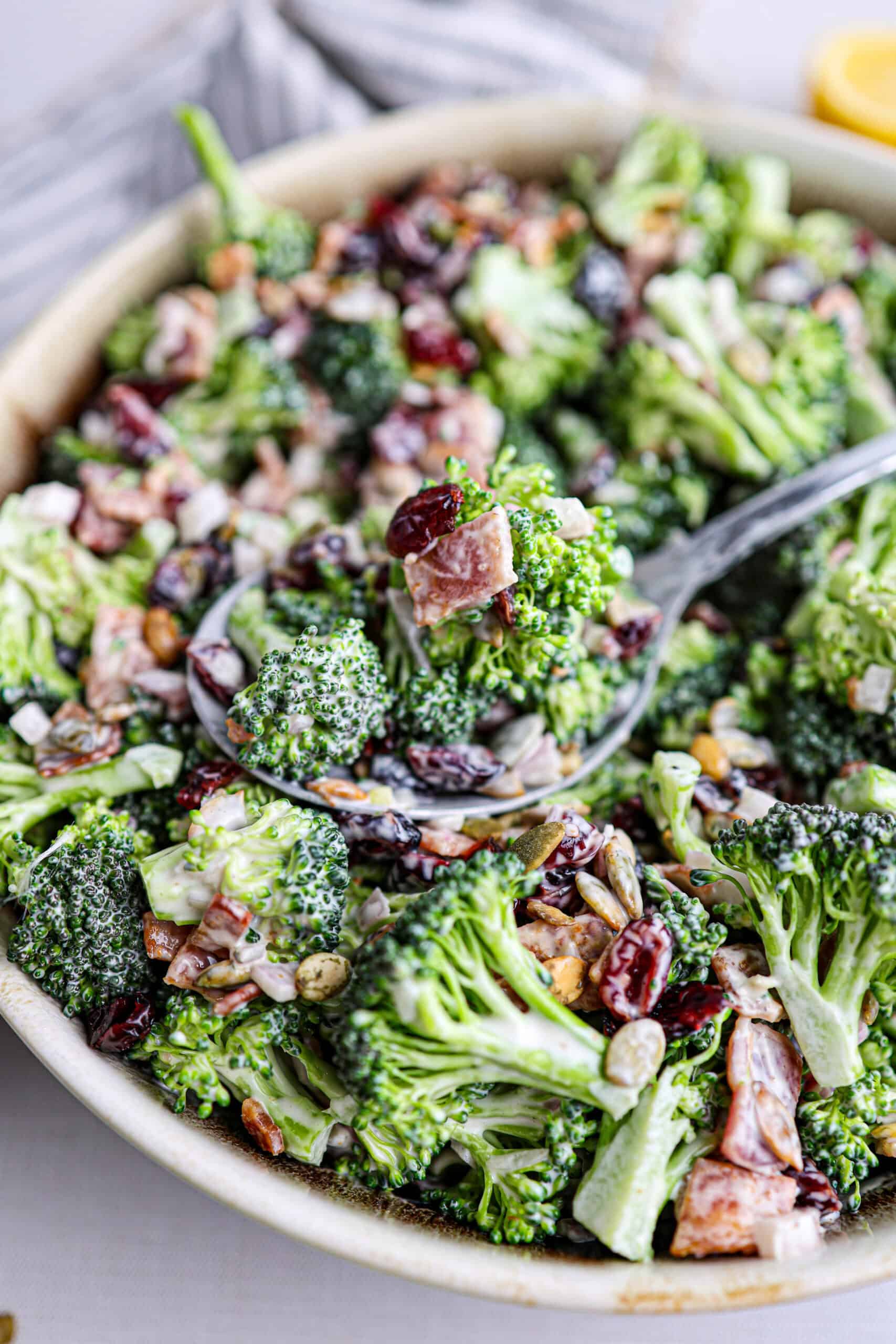 A delicious bowl of broccoli salad with bacon, dried cranberries, sunflower seeds, and a creamy dressing, served with a spoon.
