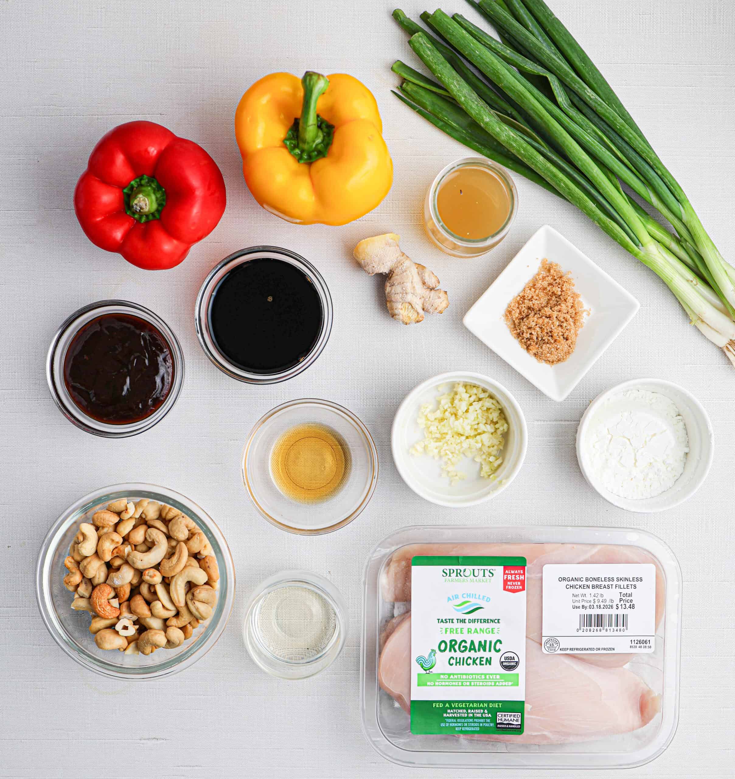 Ingredients for a cashew chicken stir-fry recipe, including bell peppers, green onions, ginger, garlic, brown sugar, cashews, sauces, and raw organic chicken breast displayed on a white surface.