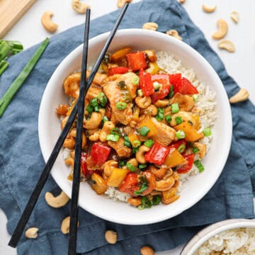 A bowl of cashew chicken stir-fried with bell peppers and rice, topped with chopped green onions and served with black chopsticks.