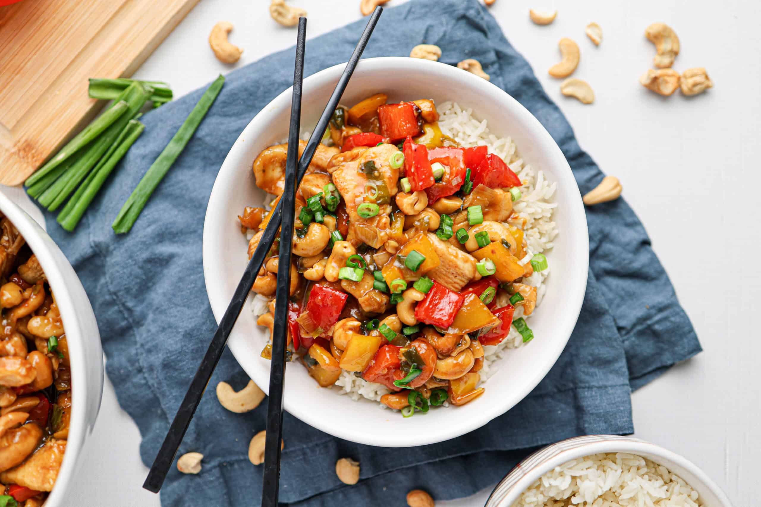 A bowl of cashew chicken stir-fried with bell peppers and rice, topped with chopped green onions and served with black chopsticks.
