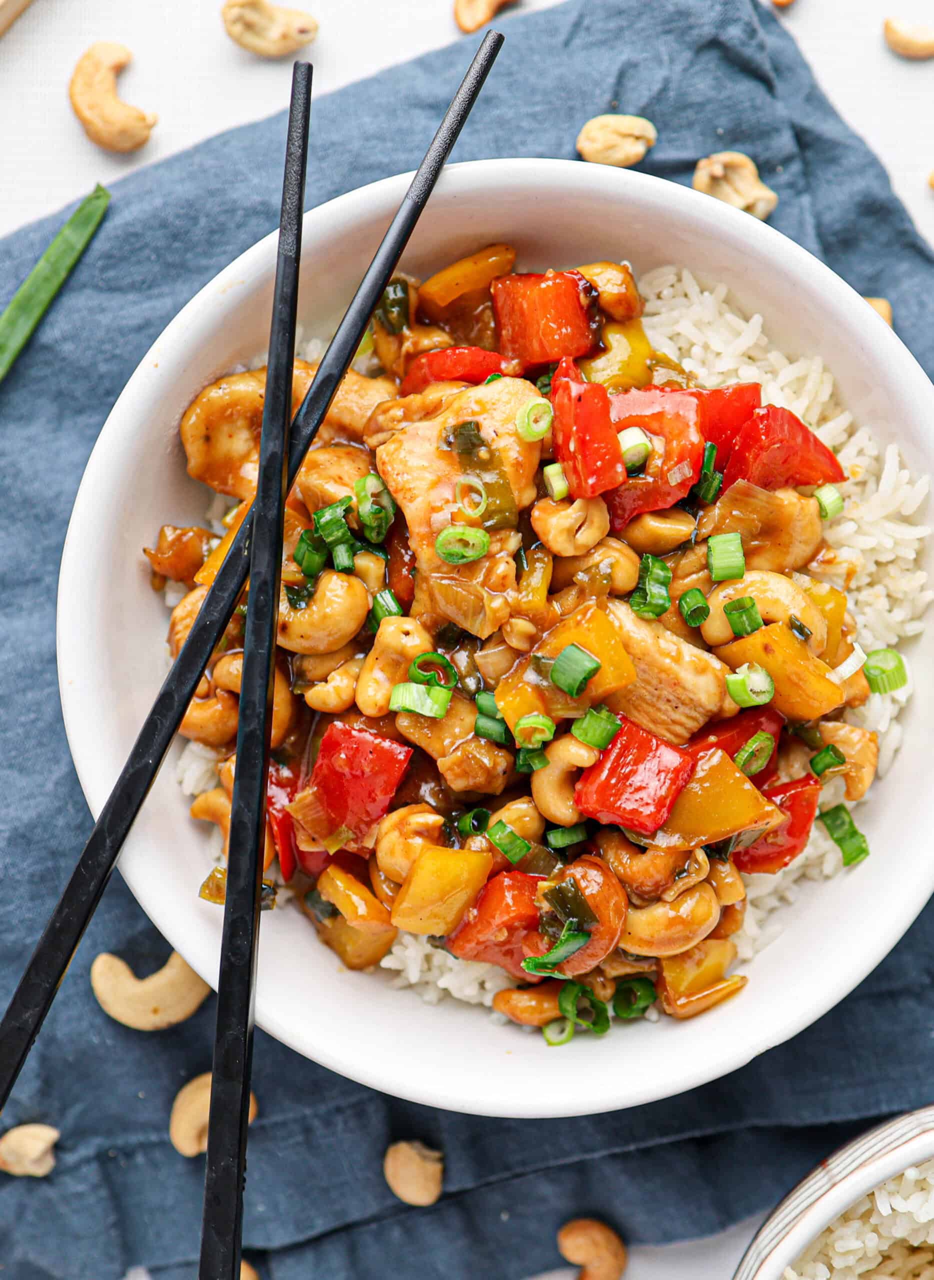 A bowl of white rice topped with savory cashew chicken, red bell peppers, cashews, and green onions, with black chopsticks resting on the bowl.