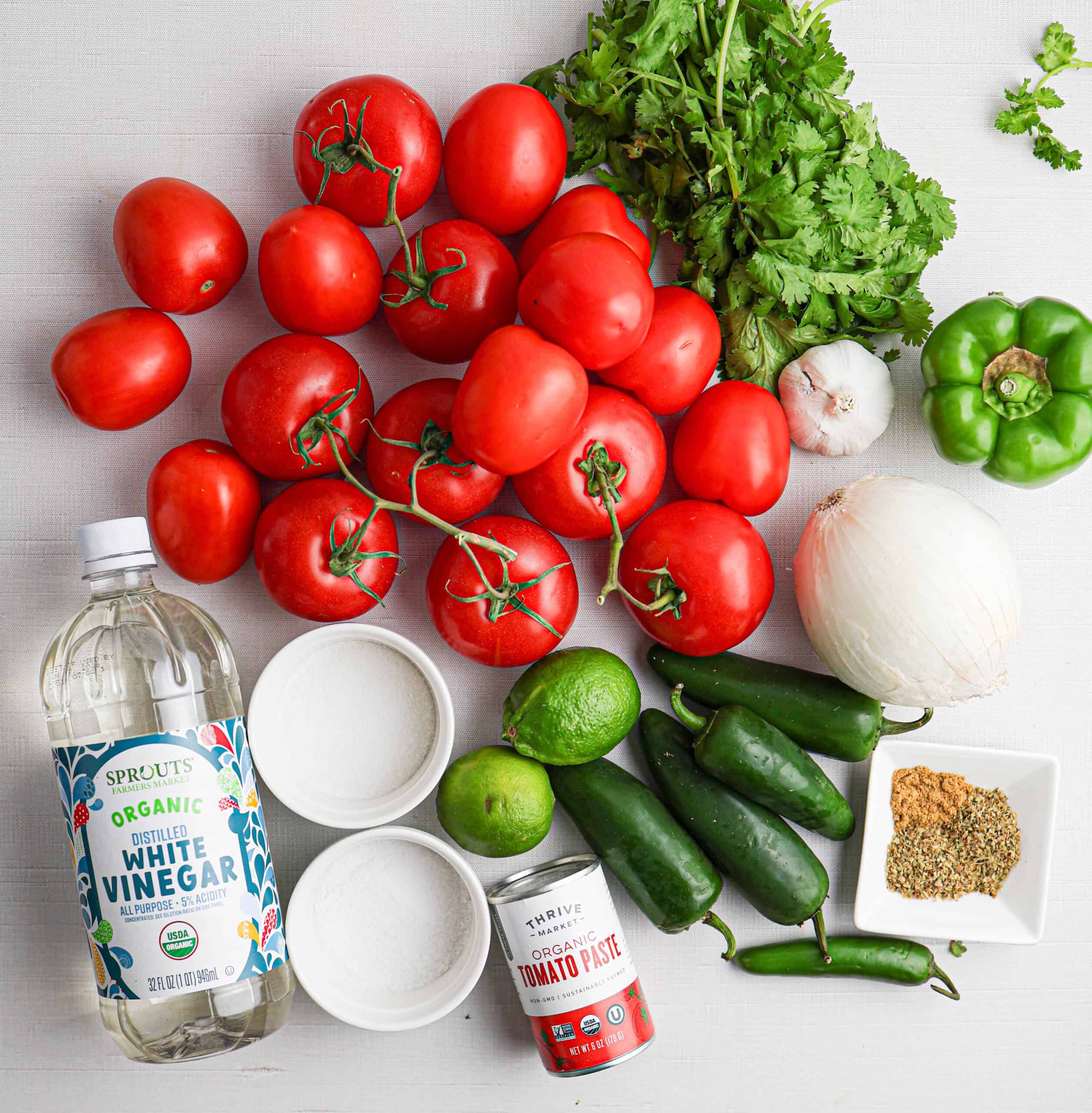 Assorted fresh tomatoes, jalapeños, green bell pepper, cilantro, white onion, lime, white vinegar, sugar, spices, and tomato paste arranged on a white surface, perfect ingredients for salsa for canning.