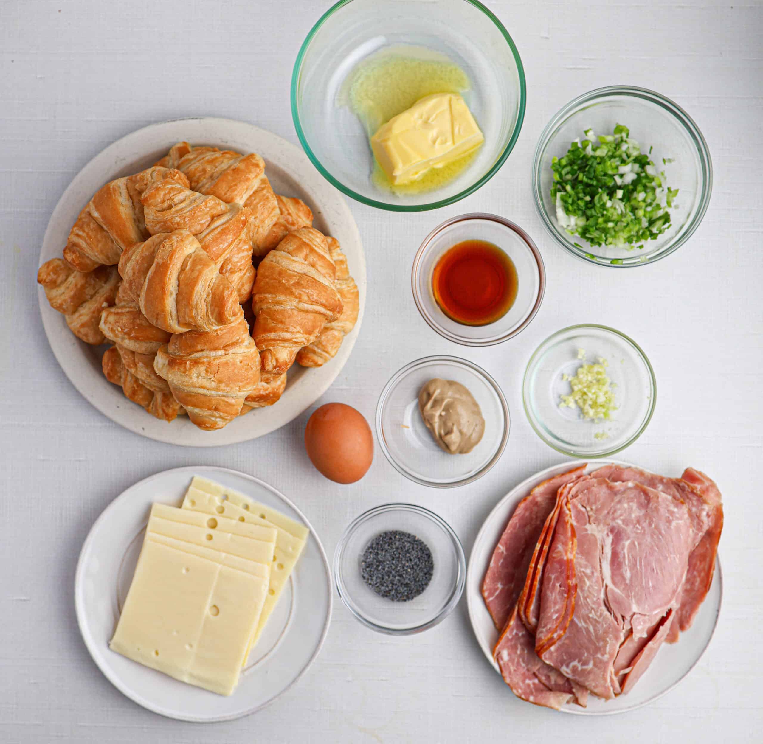 A plate of ham and cheese croissants, slices of cheese and ham, dishes of butter, chopped green onions, poppy seeds, an egg, mustard, minced garlic, and a bowl of brown liquid on a white surface.
