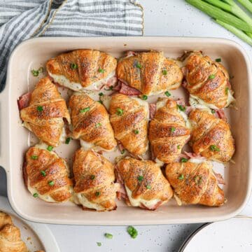 A baking dish filled with ham and cheese croissants sandwiches, topped with herbs, sits beside green onions and a striped towel.