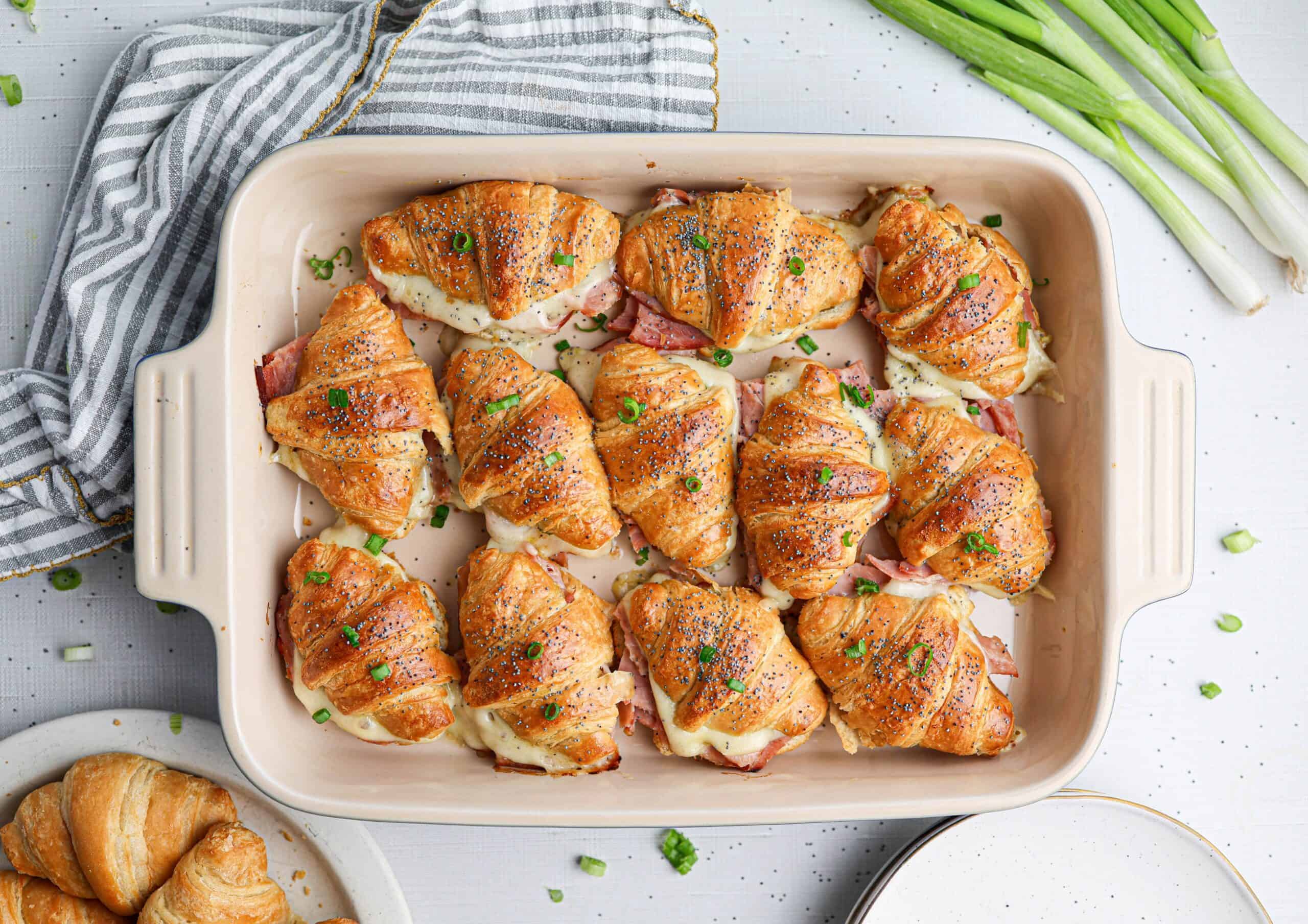 A baking dish filled with ham and cheese croissants sandwiches, topped with herbs, sits beside green onions and a striped towel.