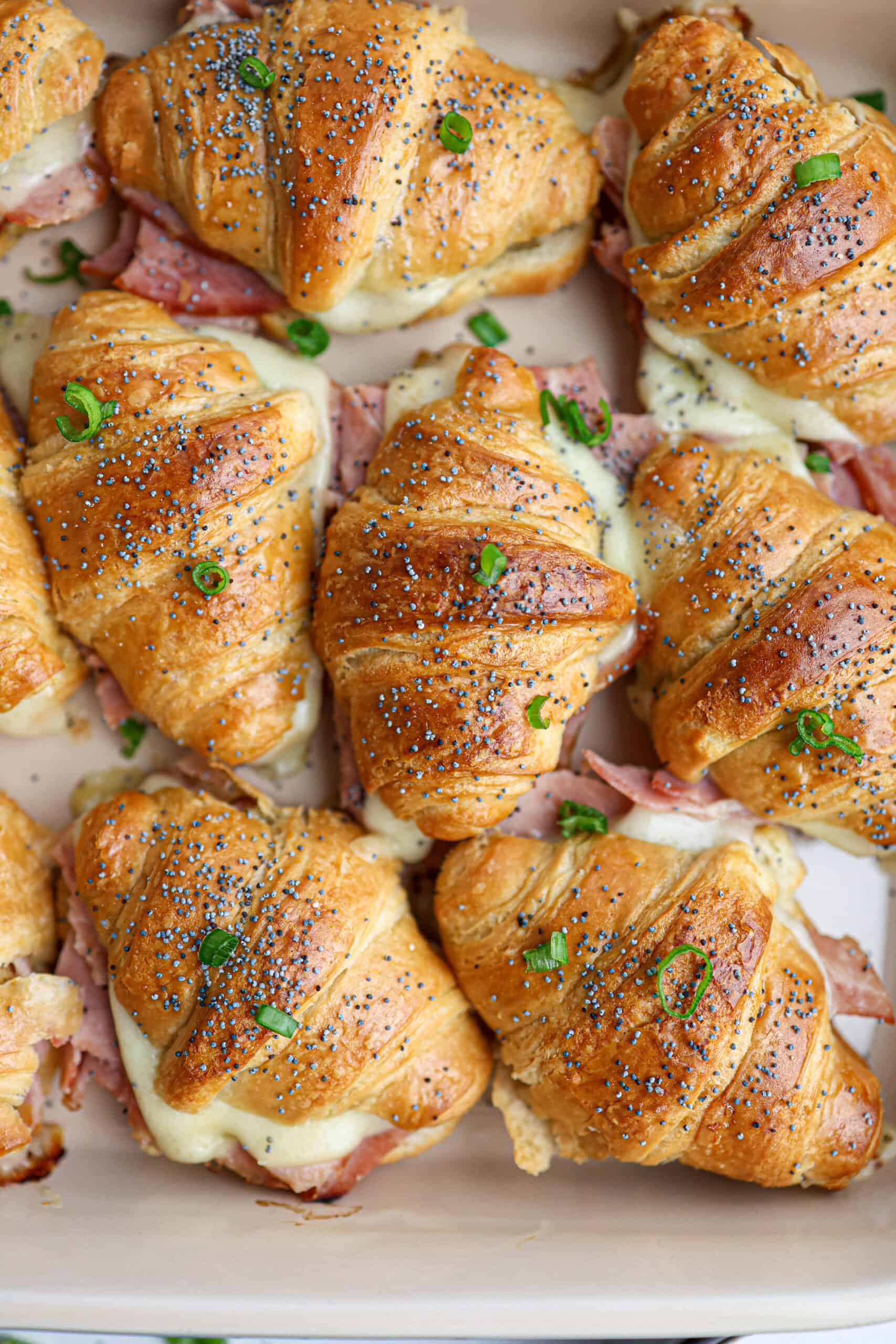 A tray of delicious ham and cheese croissants sandwiches, filled with savory ham and melted cheese, topped with poppy seeds and garnished with chopped green onions.