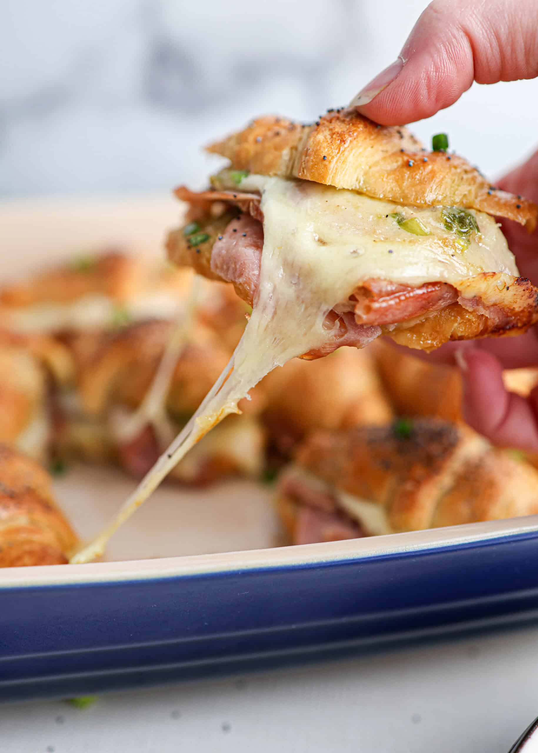 A hand lifts one of the ham and cheese croissants from a baking dish, with melted cheese stretching between the sandwich and the dish.